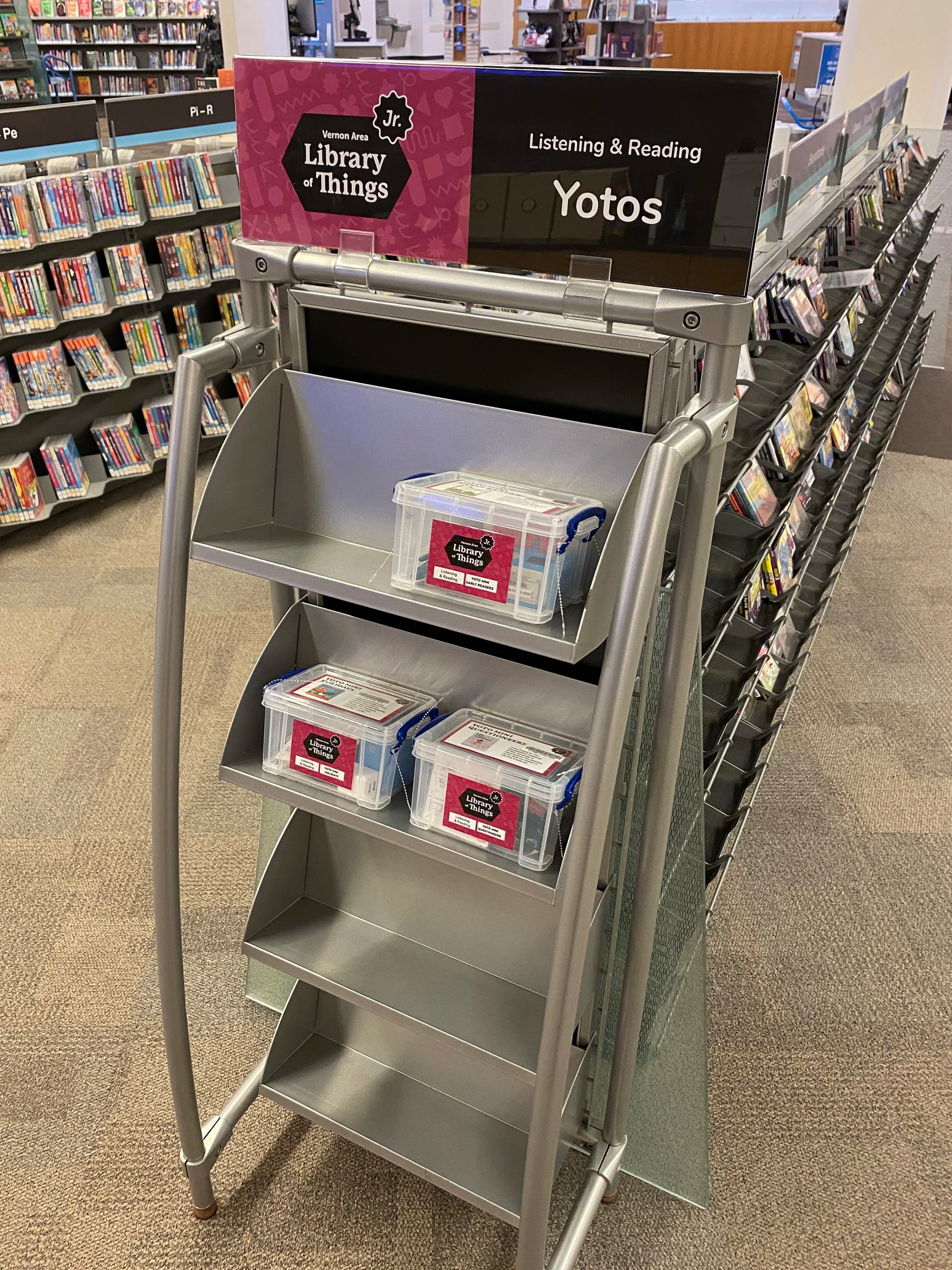A shelf display in the Vernon Area Library of Things Jr. section, holding three small clear plastic bins labeled with the program's branding, with a sign reading "Listening & Reading: Yotos."