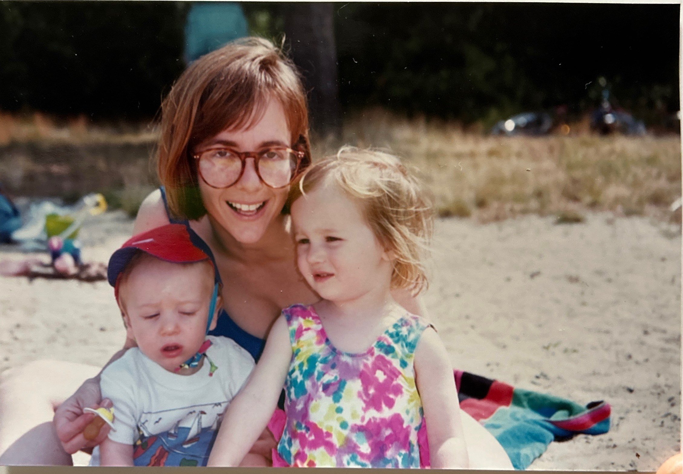 photograph of my mother, white woman with chin length light brown hair, big 80s glasses in blue bathing suit with me and my brother in front of her. i'm a little 3 year old, fair skin and blonde hair wearing floral bathing suit next to my brother