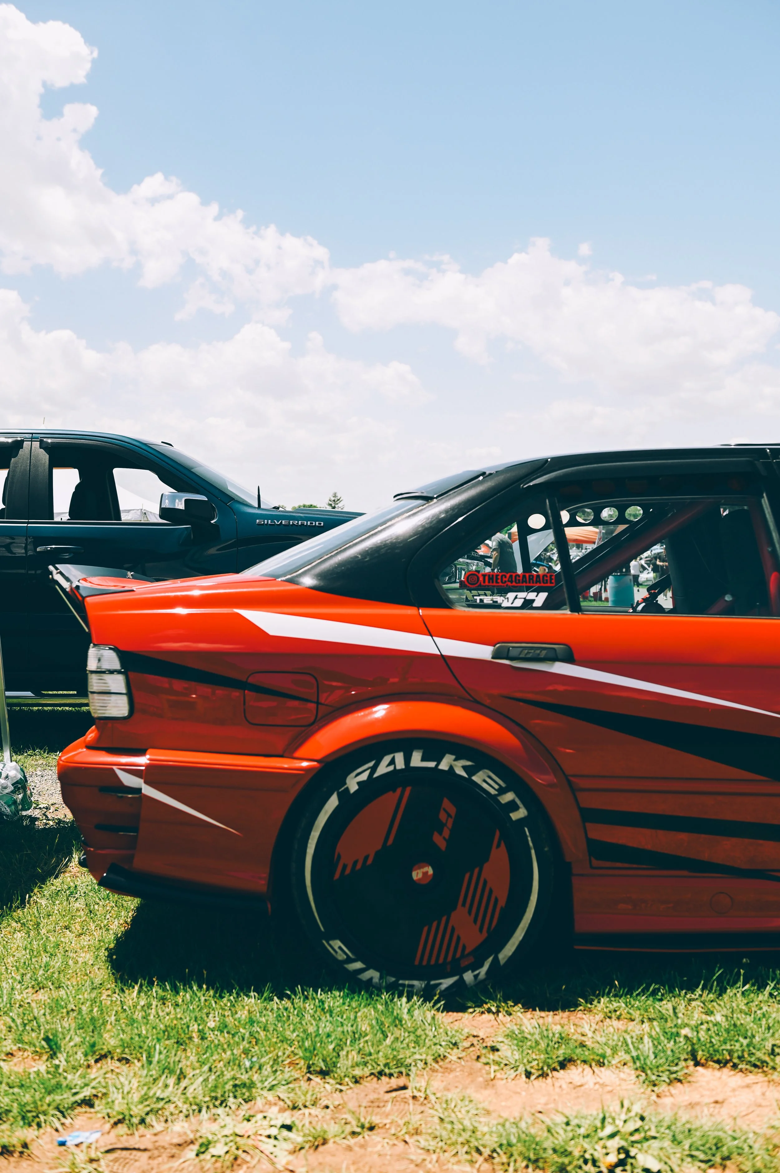 Close-up of a red race car with Falken tires parked on grass under a partly cloudy sky, with a black SUV in the background.