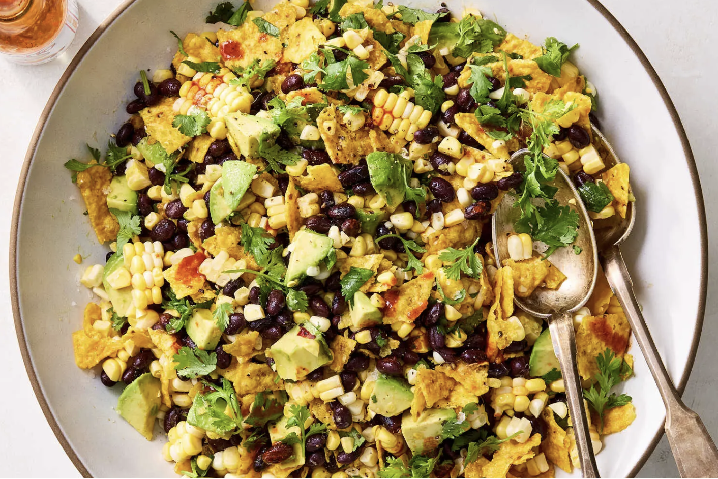 A large bowl of nachos topped with black beans, shredded cheese, avocado chunks, chopped cilantro, and olives, with serving utensils on the side.