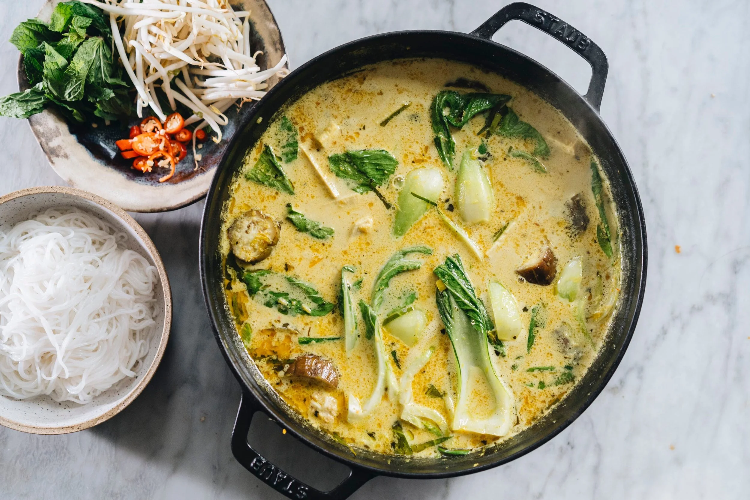 A pot of Thai green curry with vegetables, served with rice noodles and garnished with fresh herbs and chili slices.