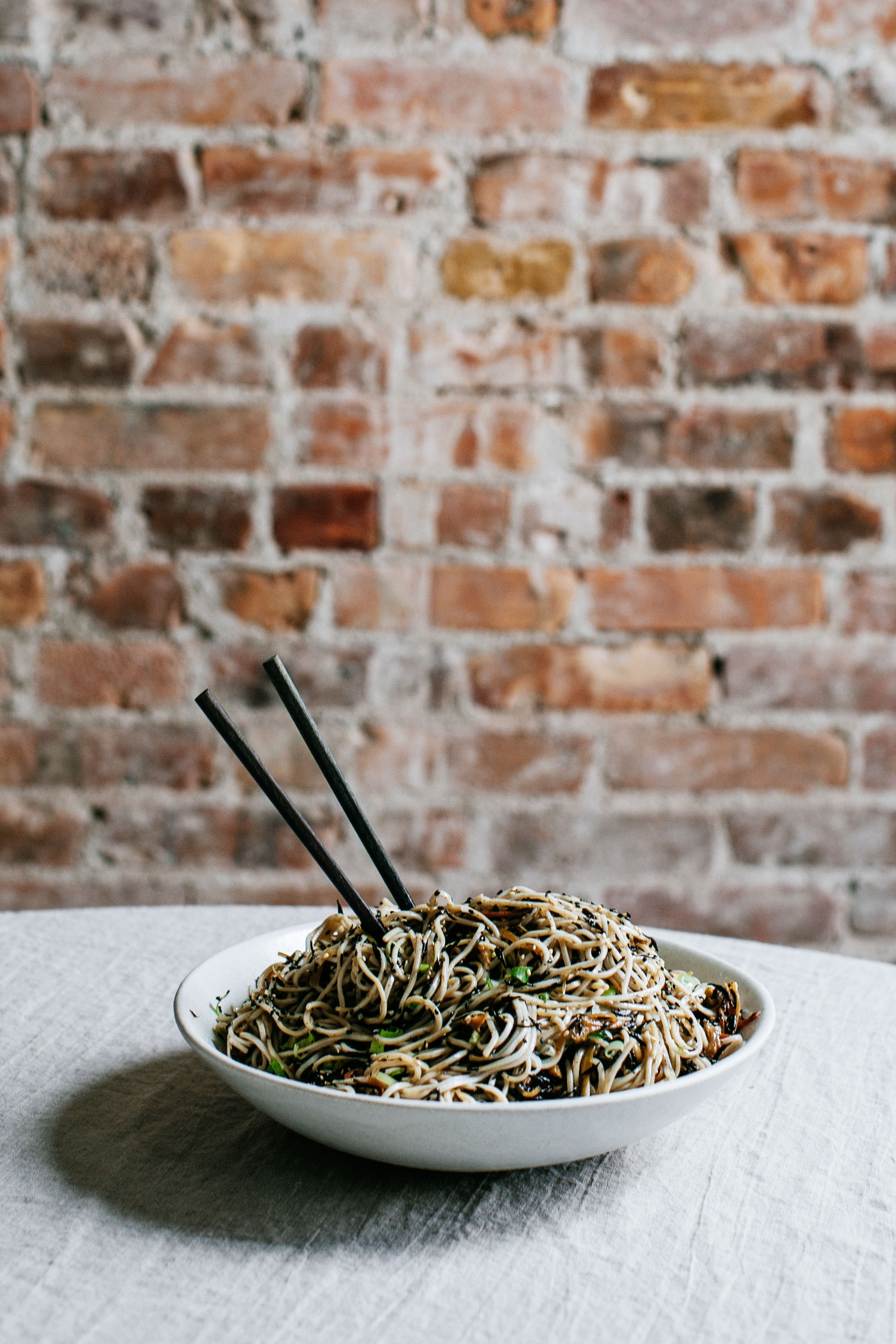 Bowl of noodles with chopsticks against brick wall backdrop.