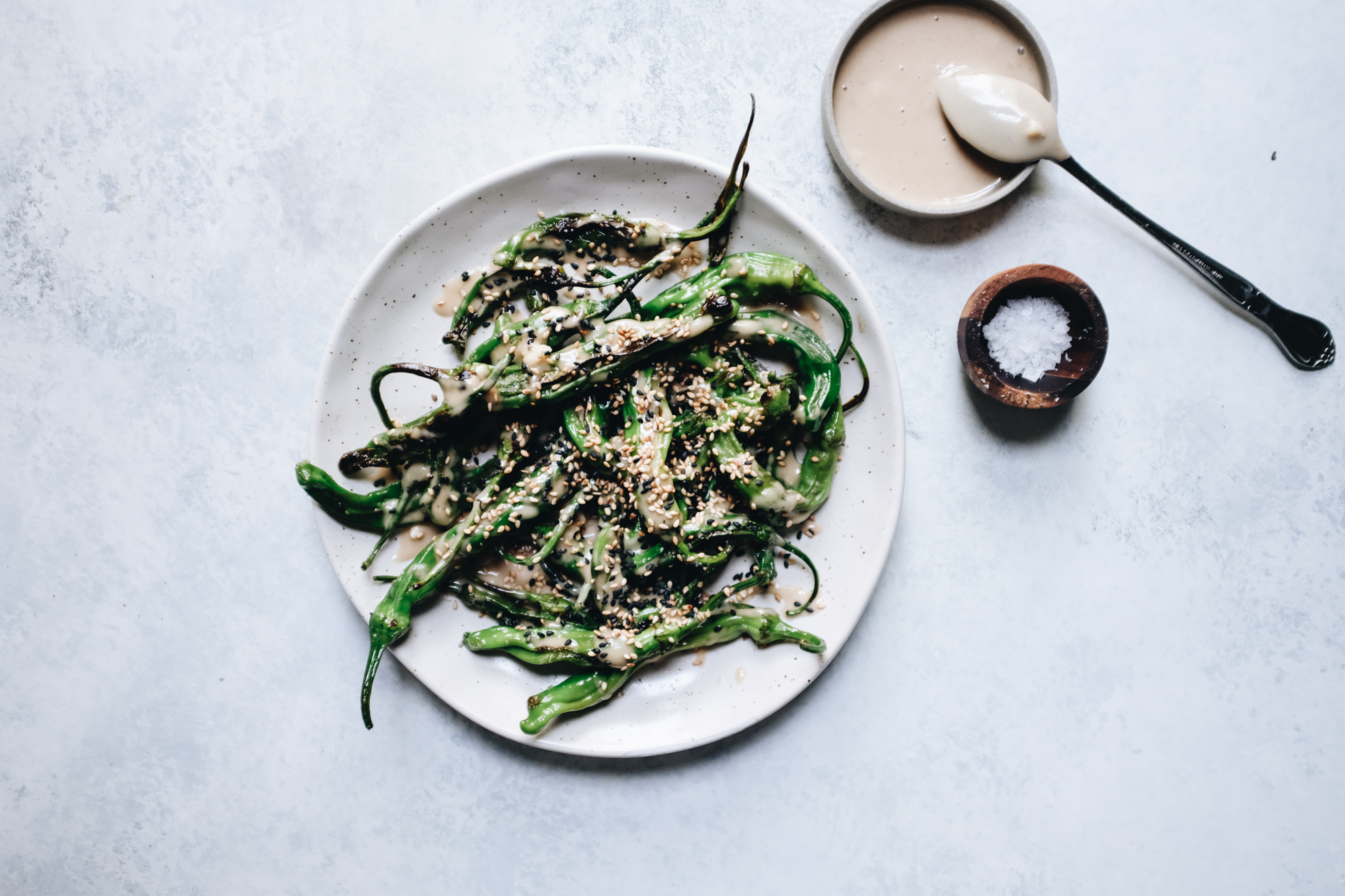 Plate of roasted shishito peppers topped with sesame seeds and sauce, next to a bowl of sauce and salt on a light background.
