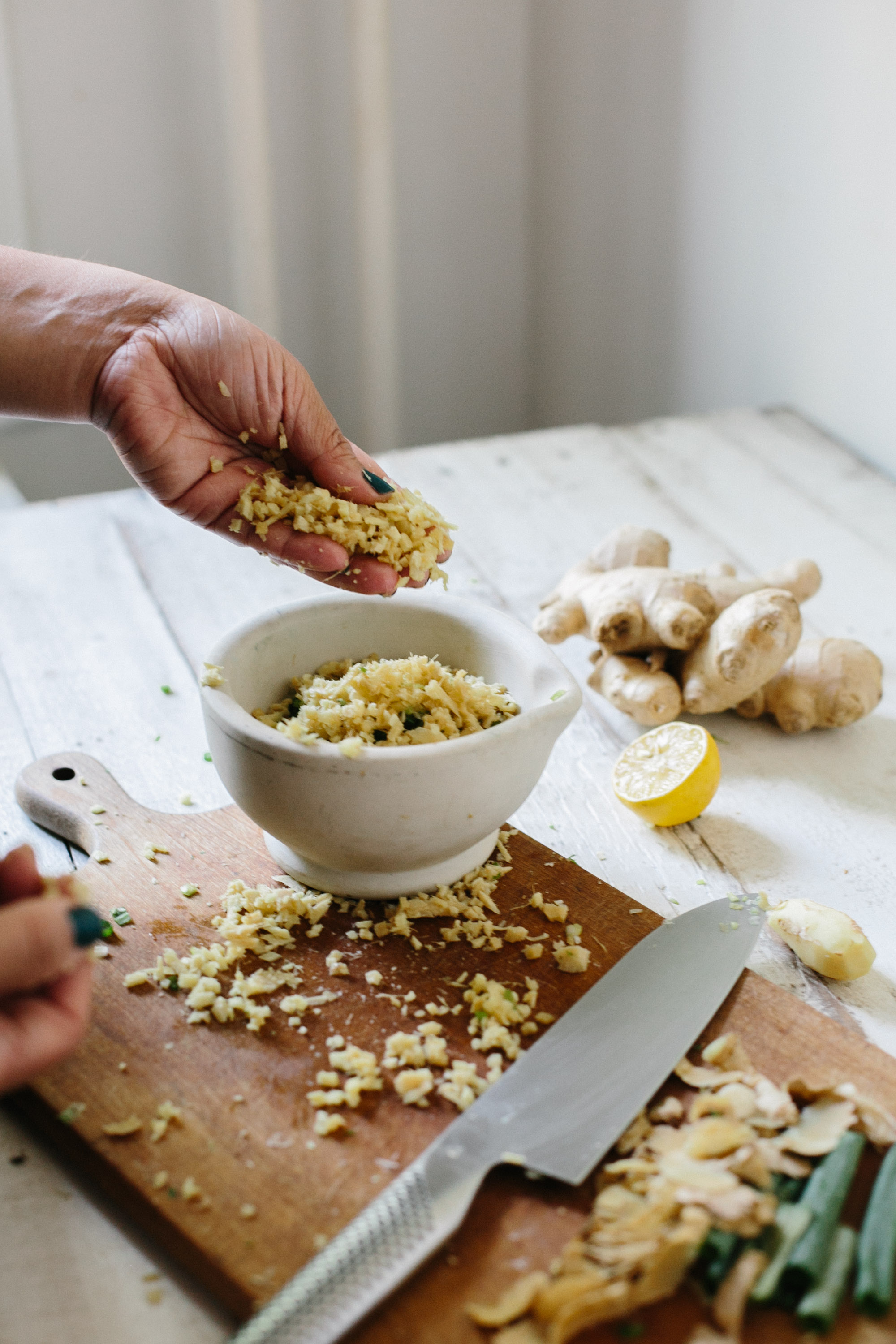 Hands preparing chopped ginger in a mortar on a wooden cutting board, with a knife, ginger root, and lemon in the background.