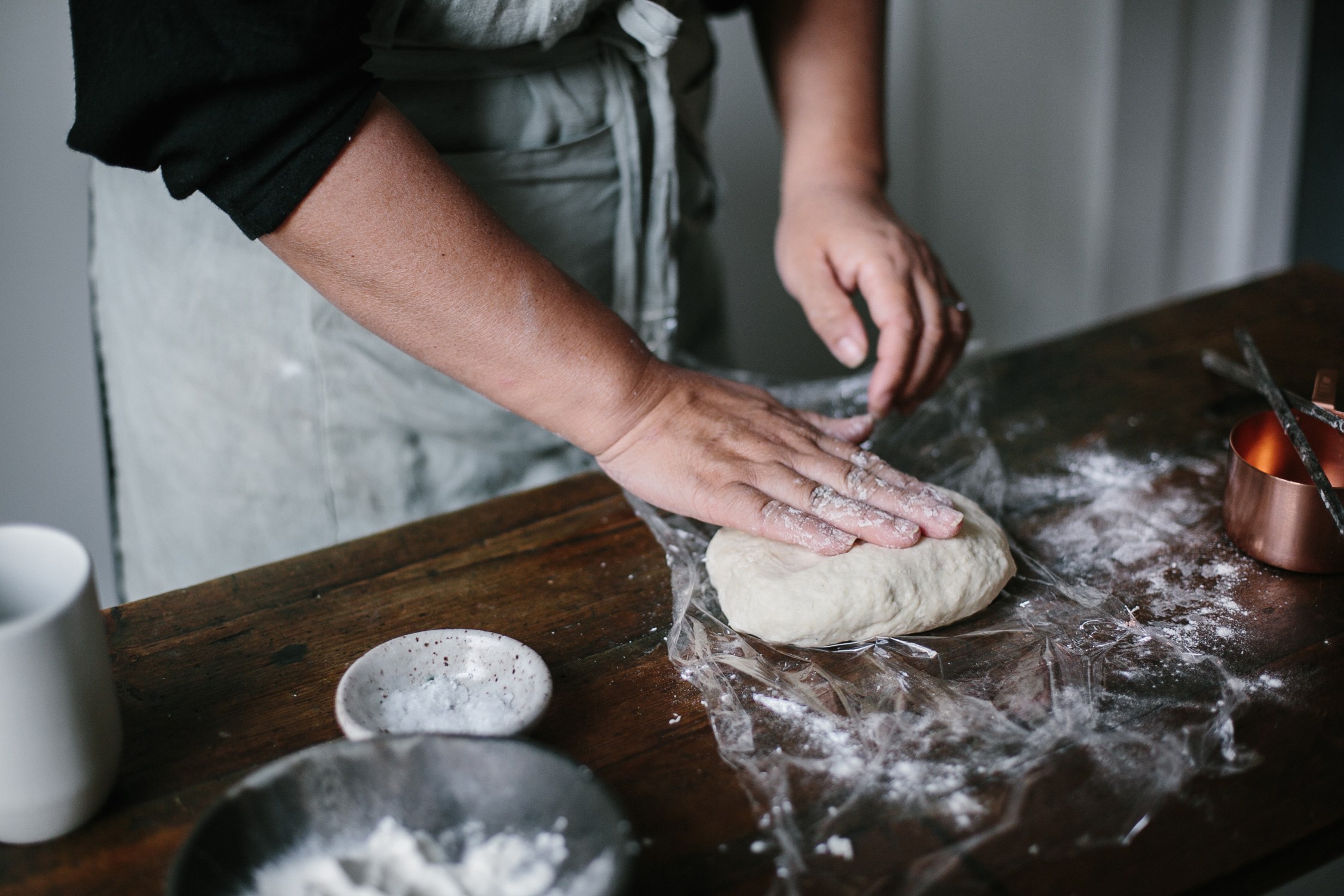 Person kneading dough on a wooden surface with flour, copper measuring cup, and small dishes.