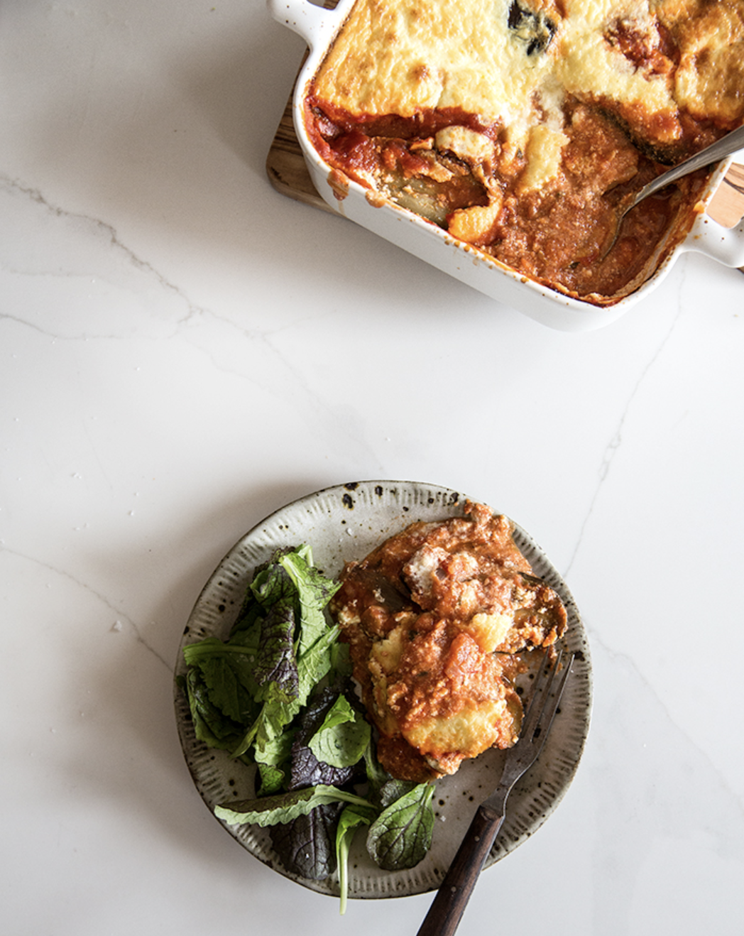 Baked lasagna in a white dish with a serving on a plate with mixed greens on a white marble surface.
