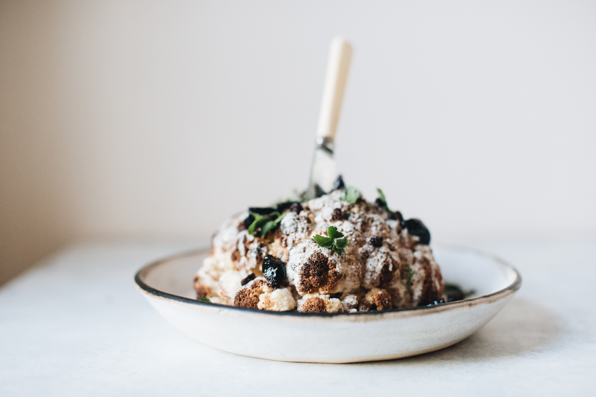 Whole roasted cauliflower garnished with herbs and spices, on a ceramic plate with a knife inserted on top.