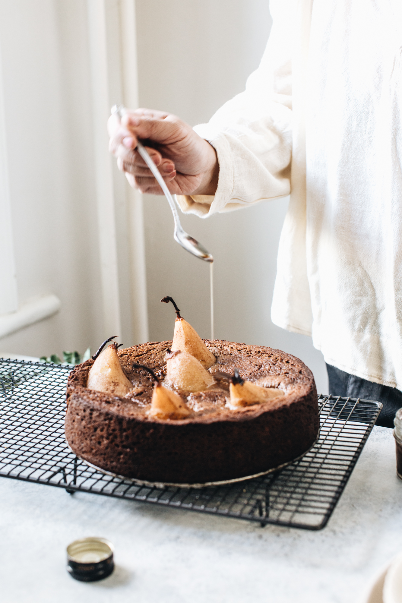 Person drizzling syrup on pear-topped chocolate cake on cooling rack.