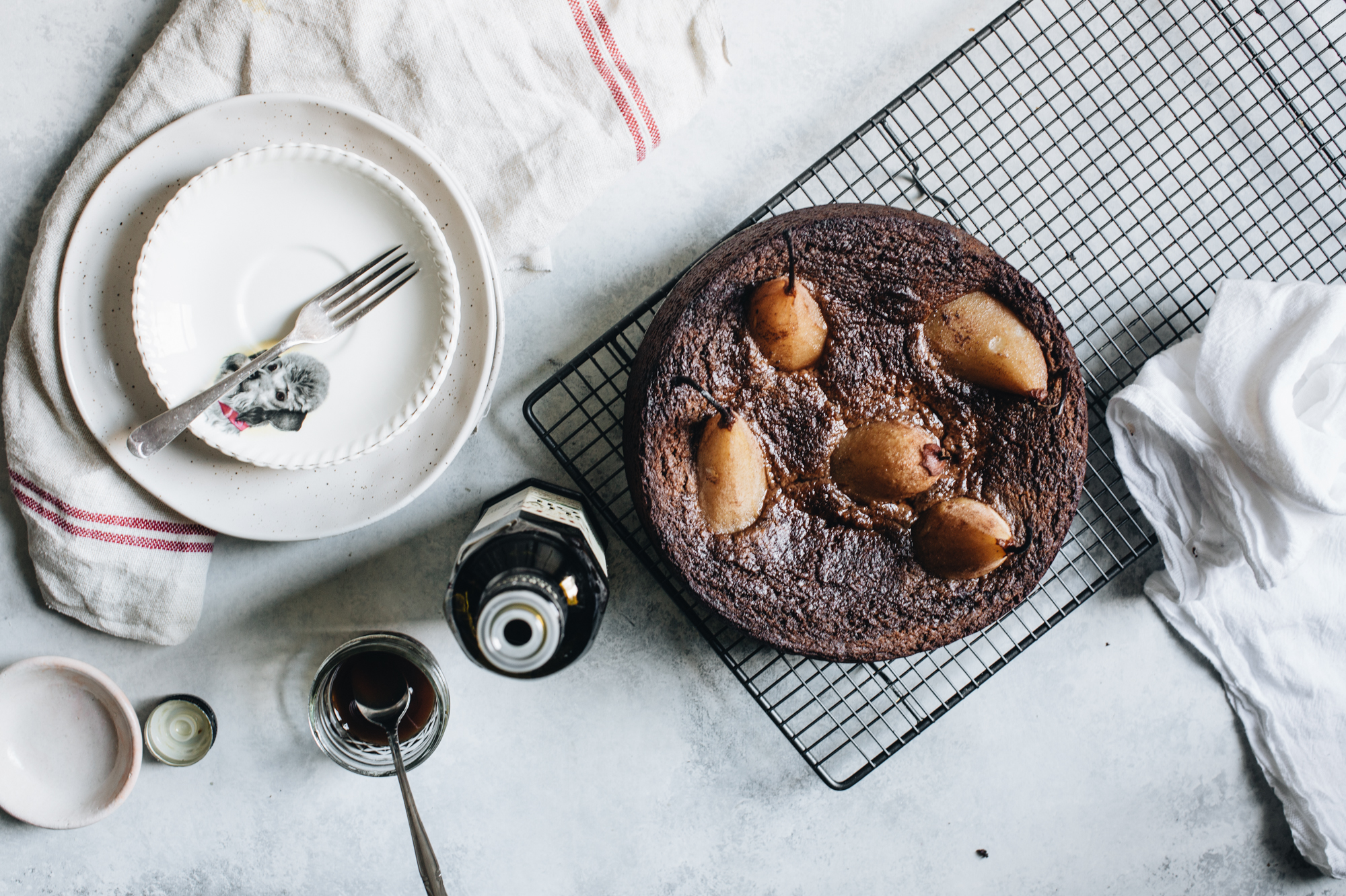 Chocolate cake with poached pears on cooling rack, surrounded by plates, fork, syrup jar, bottle, and towel on light surface.