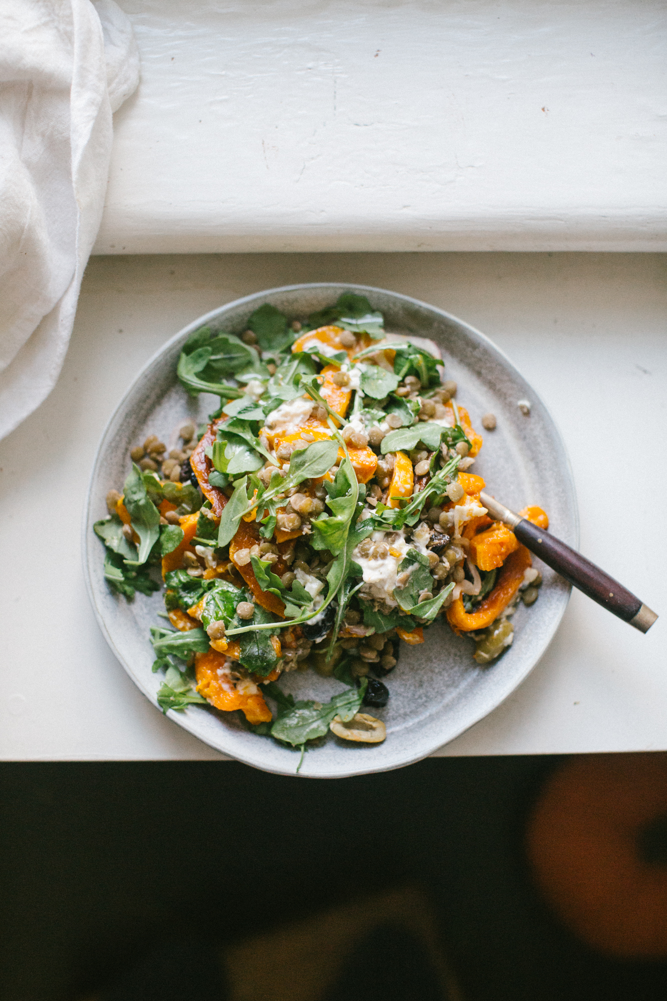 Plate of roasted vegetables, lentils, and arugula with creamy dressing on a table, fork beside plate.