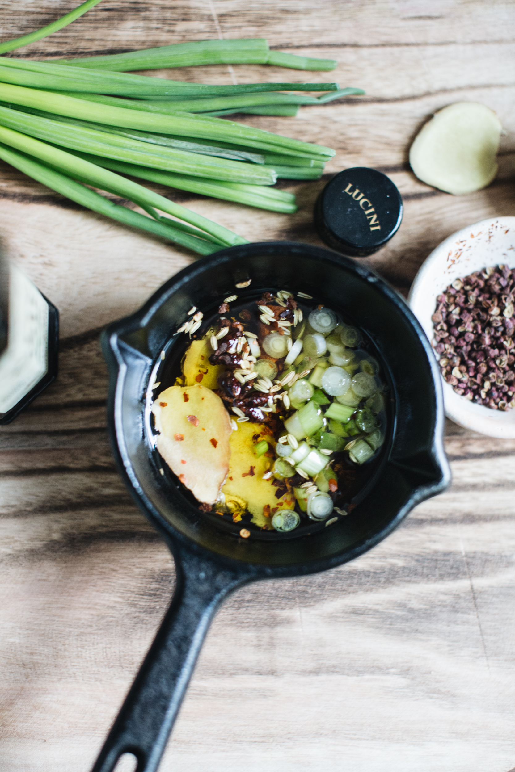 Cast iron skillet with chopped green onions, ginger slices, and spices in oil, surrounded by green onions, ginger root, and a bowl of spices on a wooden surface.