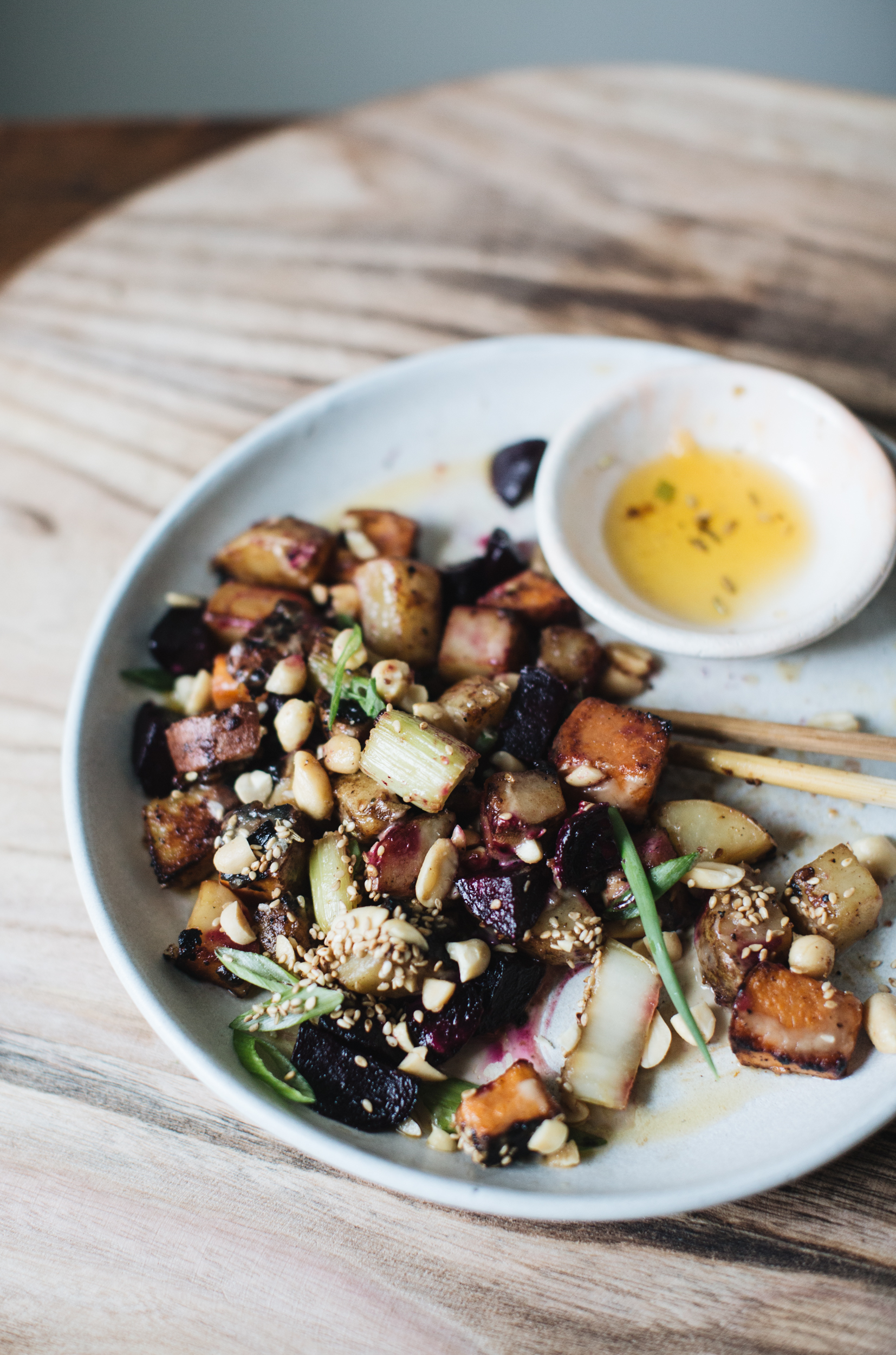 Plate of roasted root vegetables with sesame seeds and peanuts, small bowl of dipping sauce.