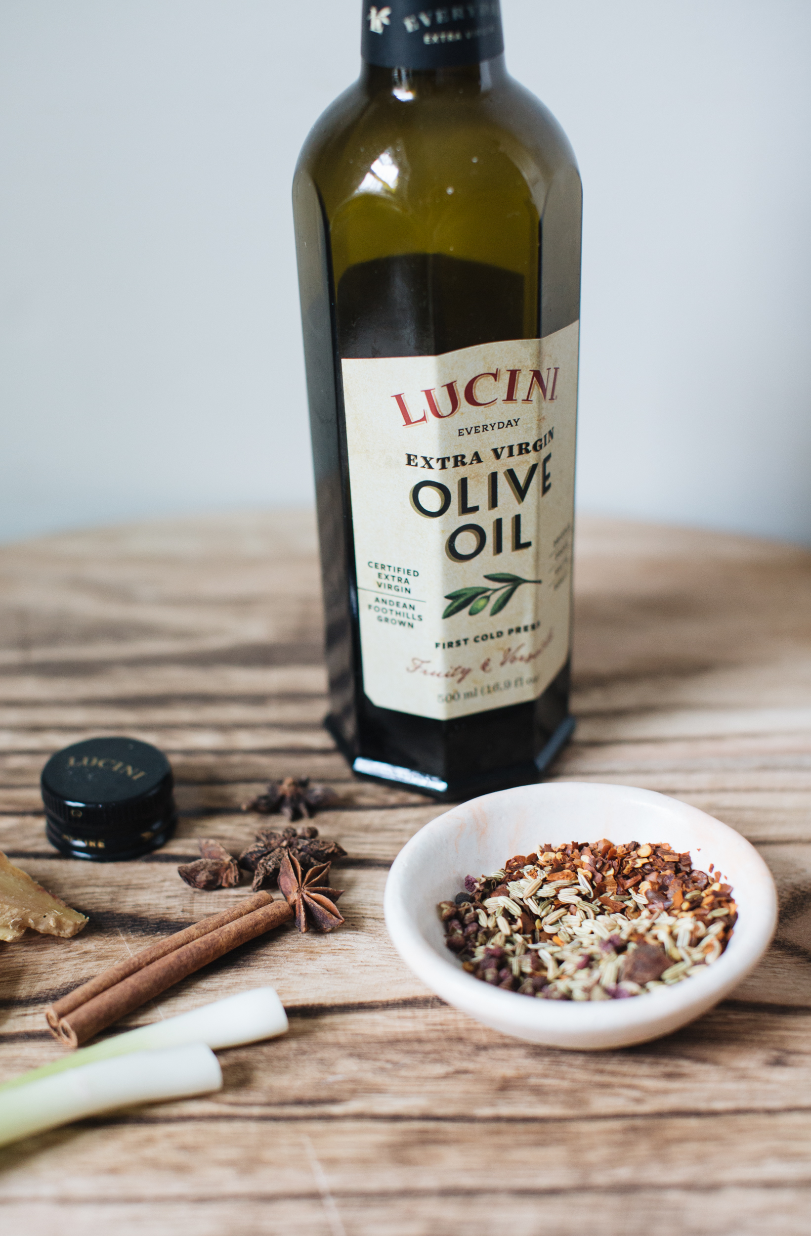 Bottle of Lucini Extra Virgin Olive Oil on a wooden table with a bowl of mixed spices, star anise, cinnamon sticks, and a piece of ginger.