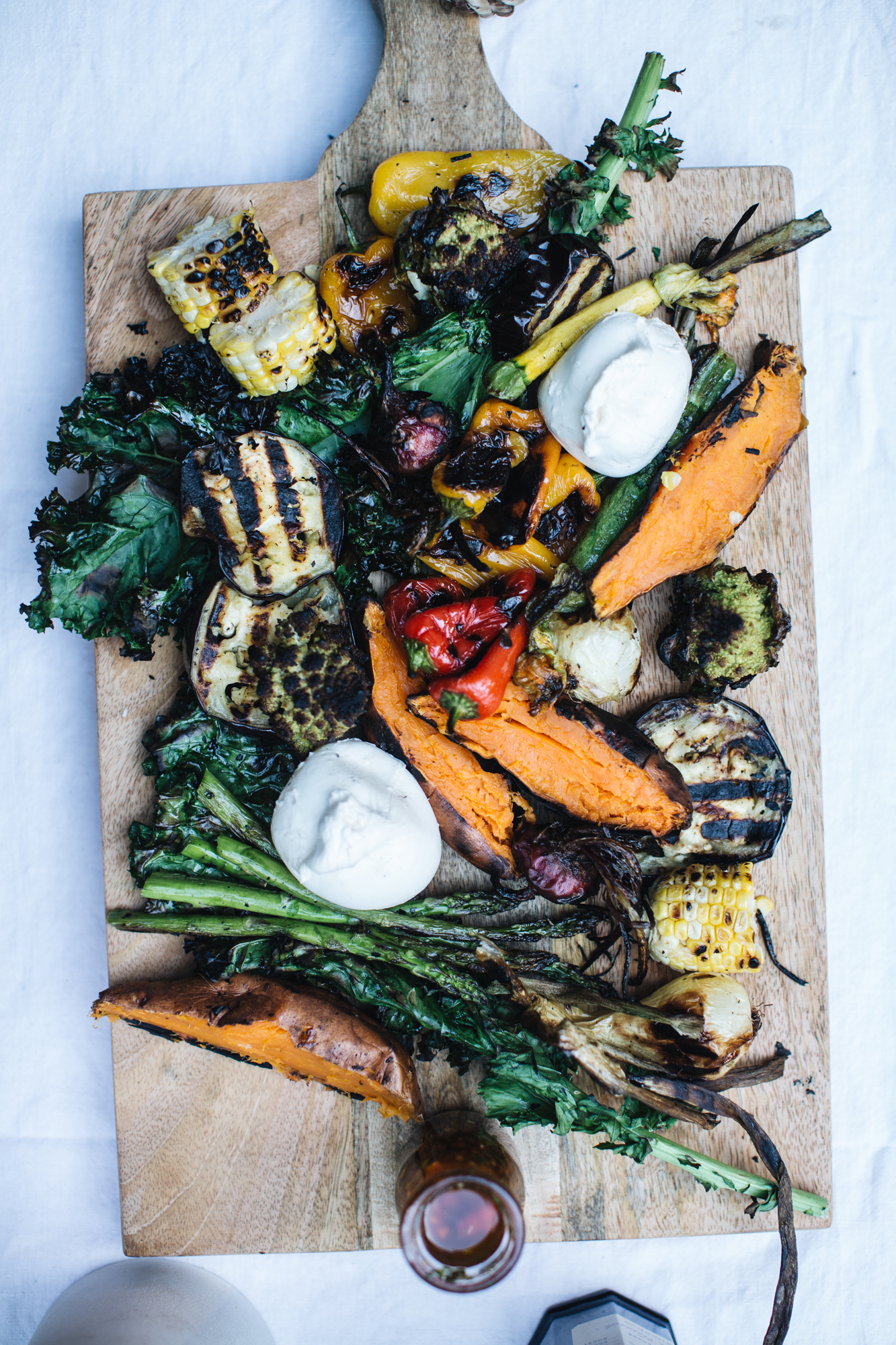 Grilled vegetables on a wooden board, including sweet potatoes, corn, eggplant, asparagus, kale, bell peppers, and dollops of cheese.