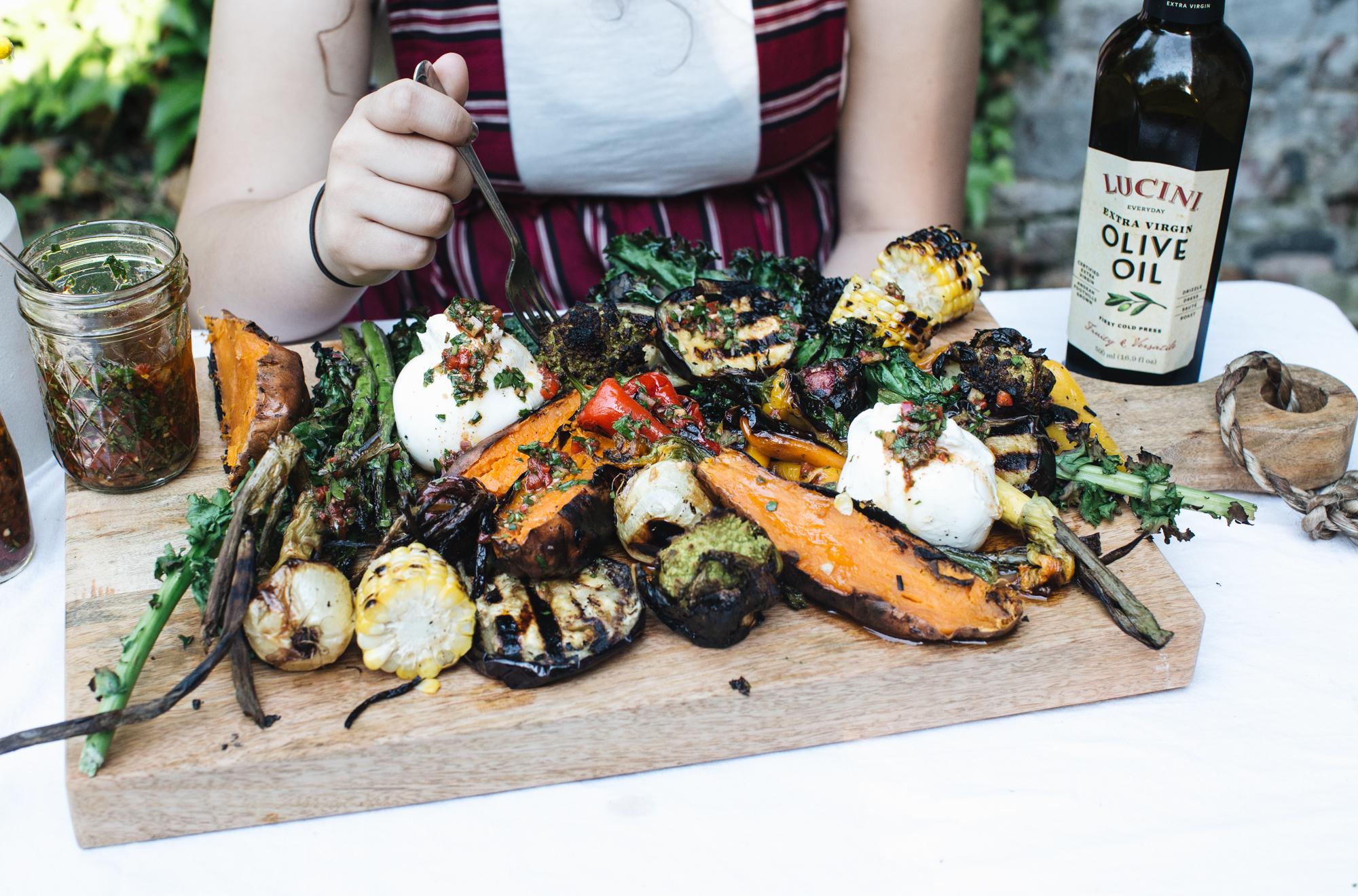 A person eating a meal featuring grilled vegetables such as corn, peppers, and sweet potatoes, topped with fresh mozzarella and herbs. A jar of sauce and a bottle of olive oil are also on the wooden serving board.