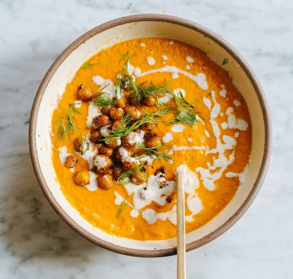 A bowl of creamy orange-colored soup garnished with roasted chickpeas, fresh herbs, and a drizzle of cream on top, with a spoon resting inside.