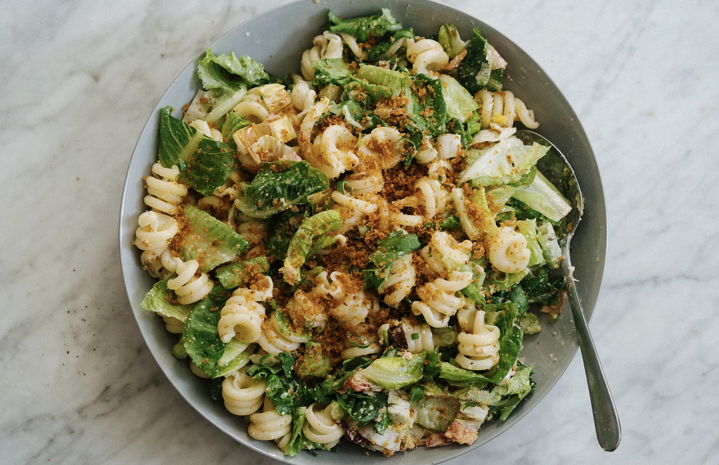 Bowl of pasta salad with rotini, lettuce, and seasoning, on a marble surface with a spoon.