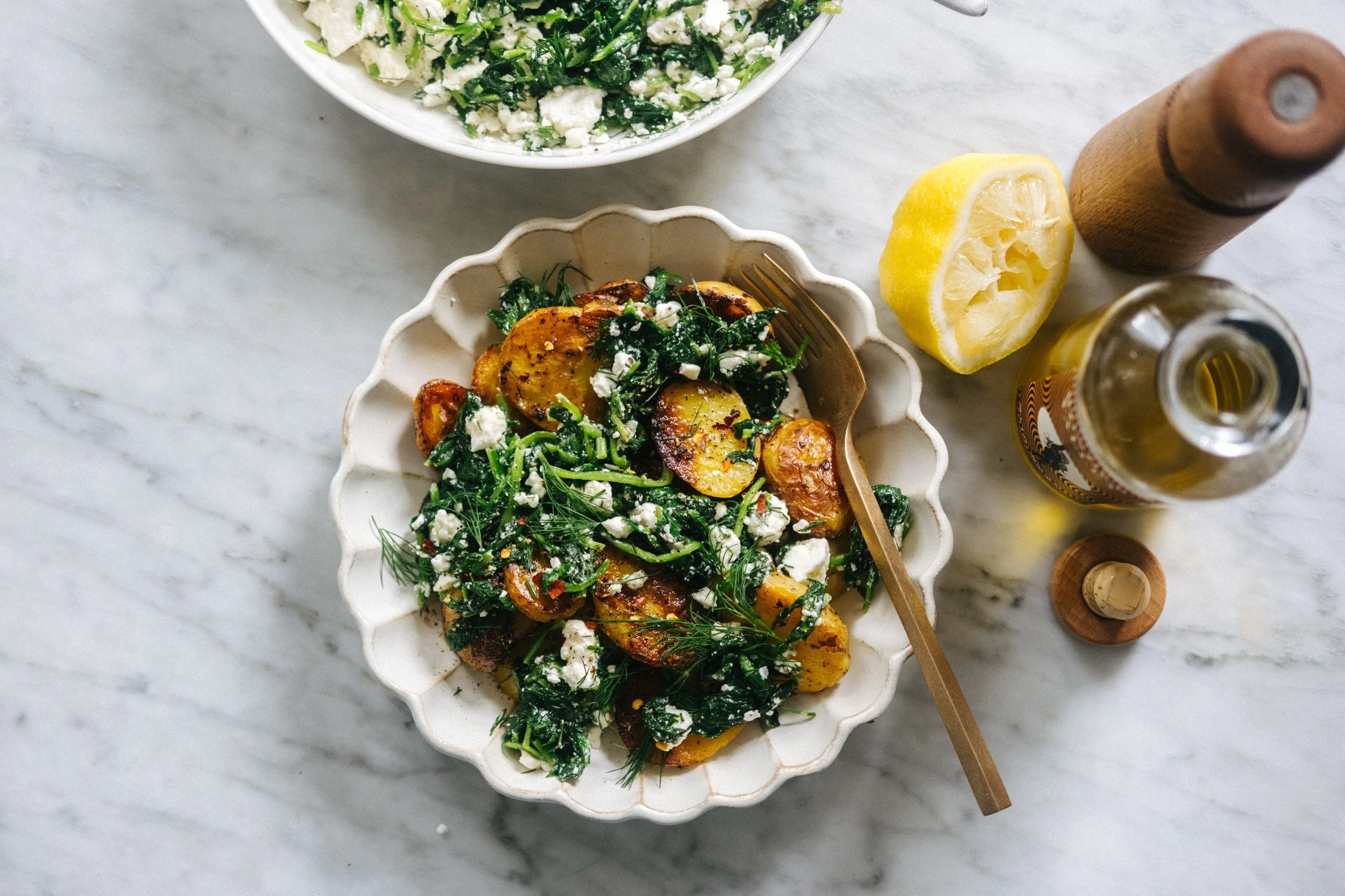 A bowl of roasted potatoes topped with herbs and feta cheese, accompanied by a lemon half, a bottle of oil, and a pepper grinder on a white marble surface.