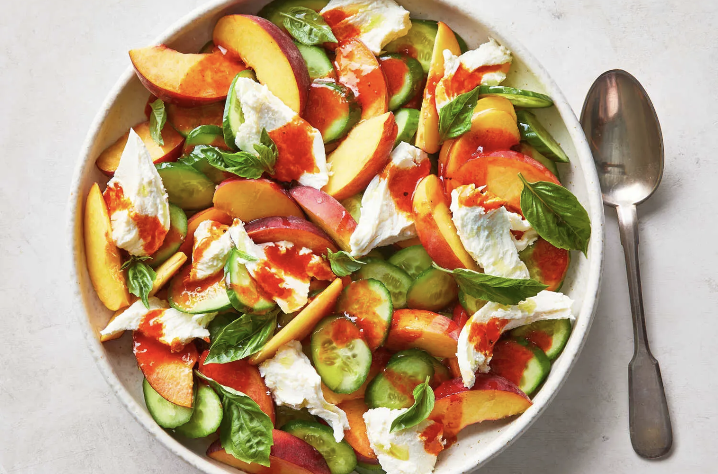 Peach and cucumber salad with feta cheese, basil leaves, and red dressing in a white bowl with a spoon