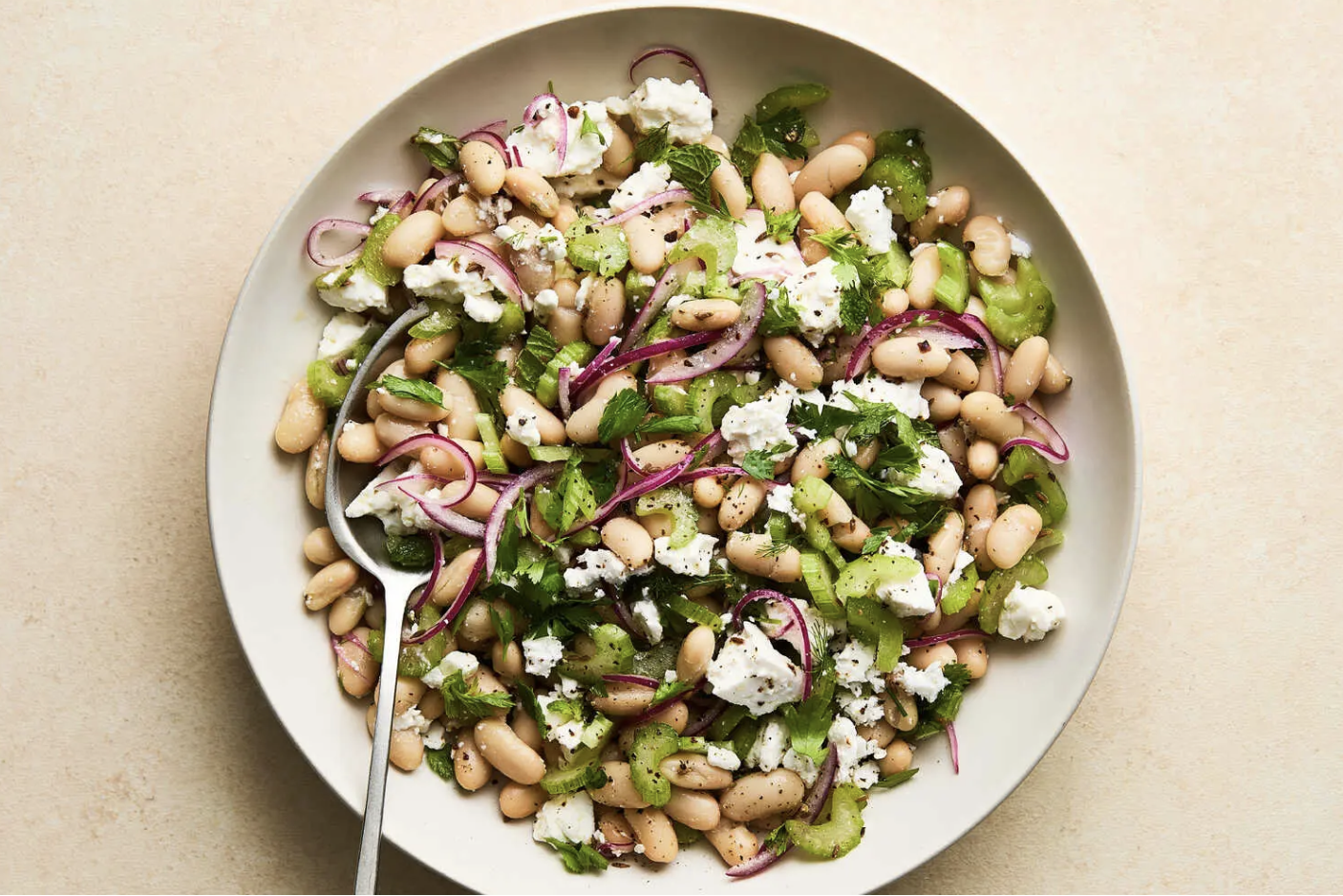 White bean salad with red onion, celery, herbs, and feta cheese in a white bowl.