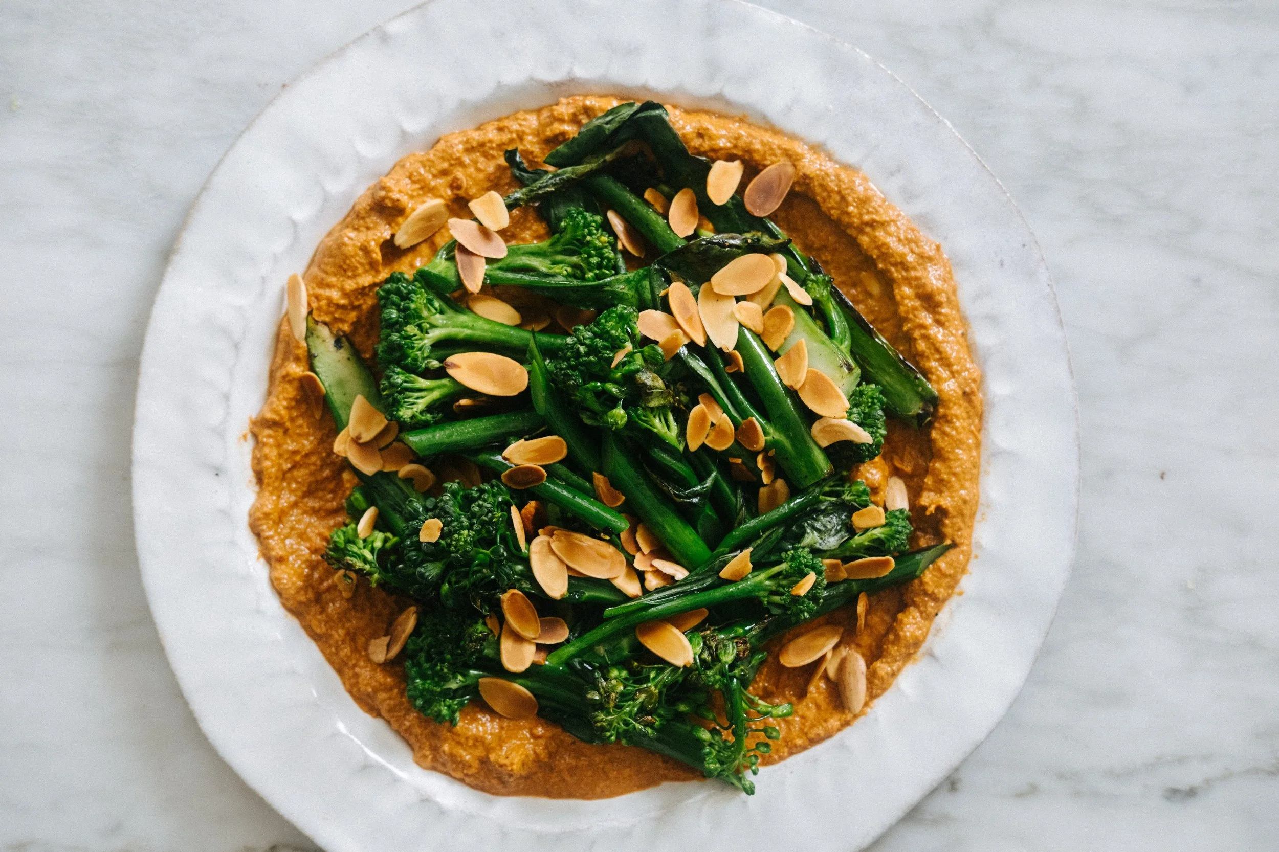 A white plate holding a dish with sautéed green broccoli and spinach topped with sliced almonds, served on a white marble surface.