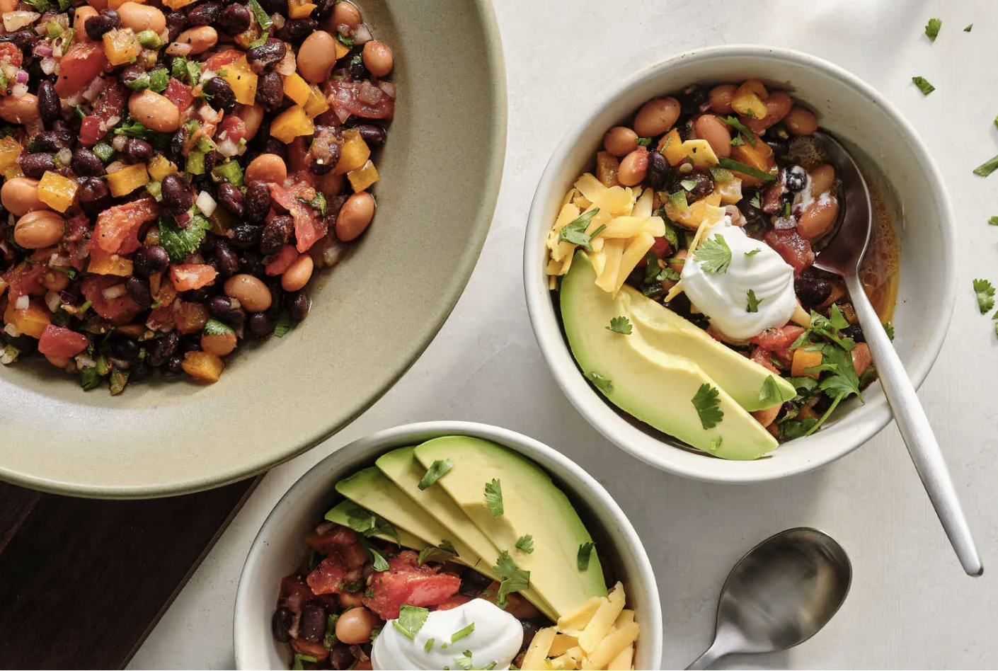 Two bowls of black bean and vegetable chili garnished with avocado slices, shredded cheese, sour cream, and cilantro, with a larger bowl of the bean and vegetable mixture in the background.