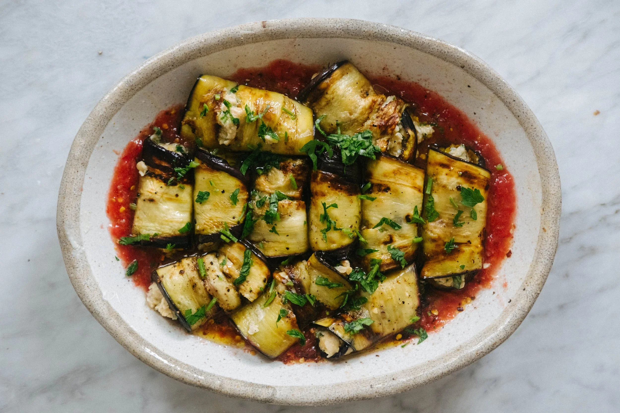 Grilled zucchini pieces topped with chopped parsley in a red sauce in a beige bowl on a marble surface.