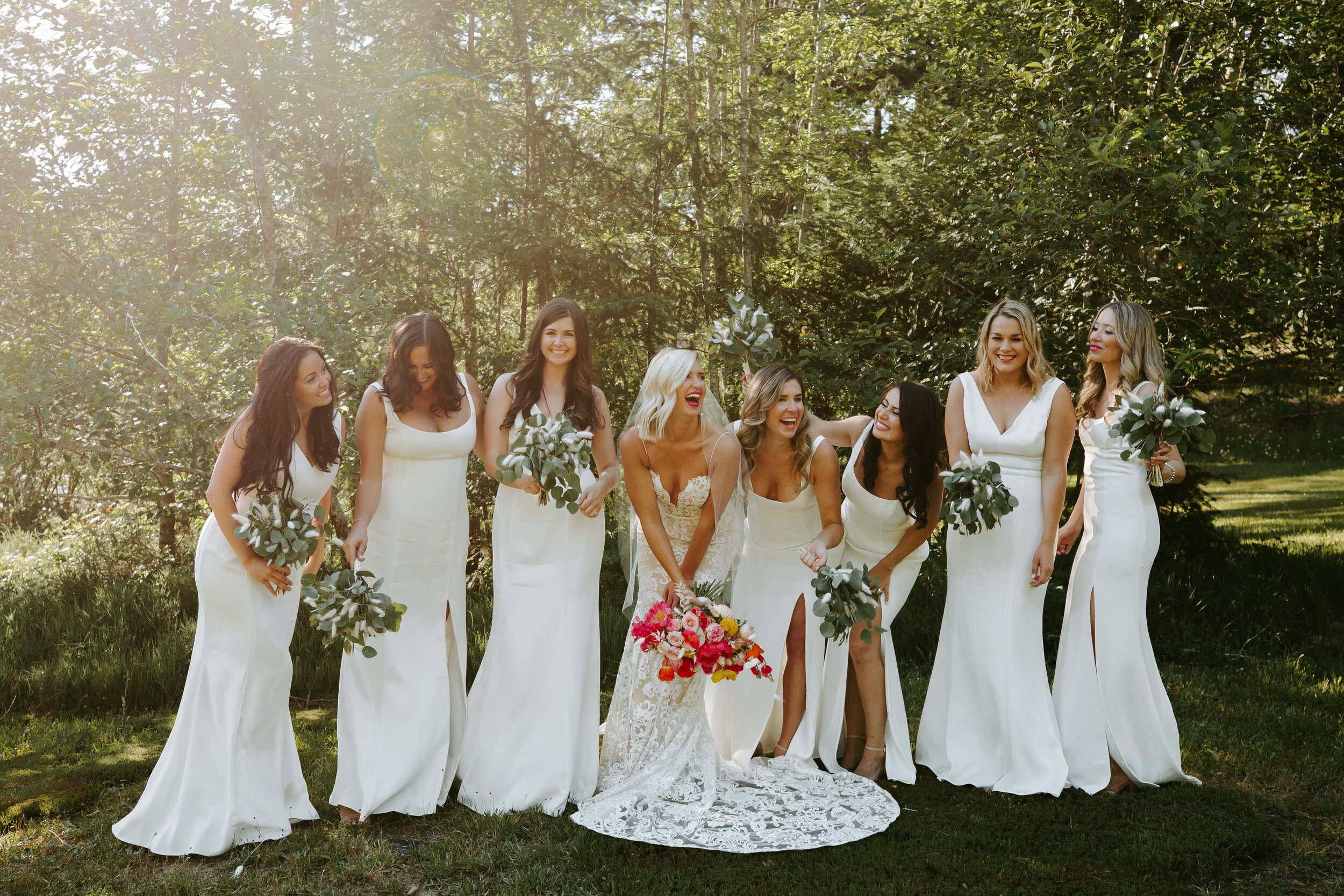 Group of women in white dresses outdoors, smiling and holding bouquets, celebrating a wedding.
