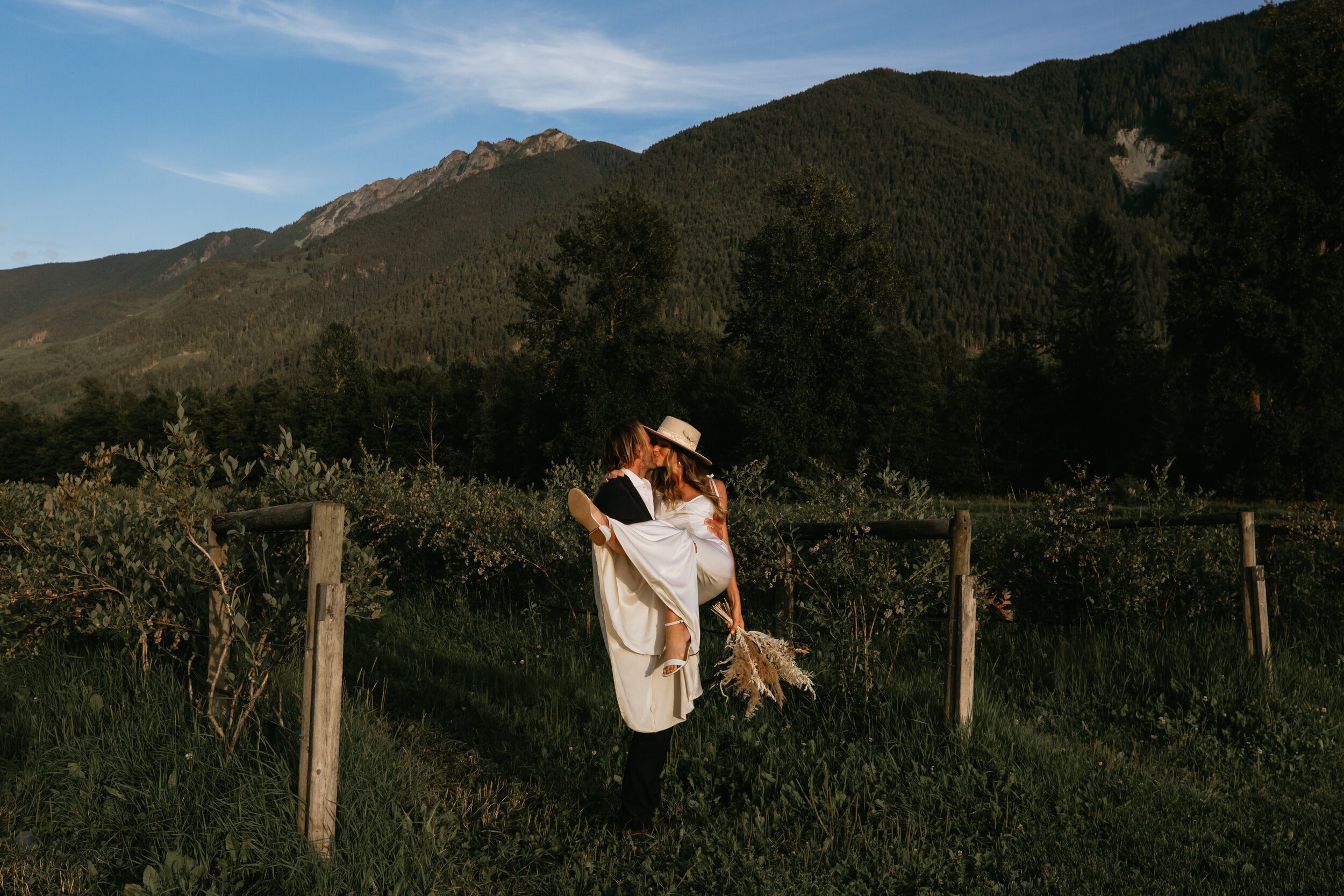 A couple dressed in wedding attire standing on a grassy field, sharing a kiss, with mountains and trees in the background during sunset.