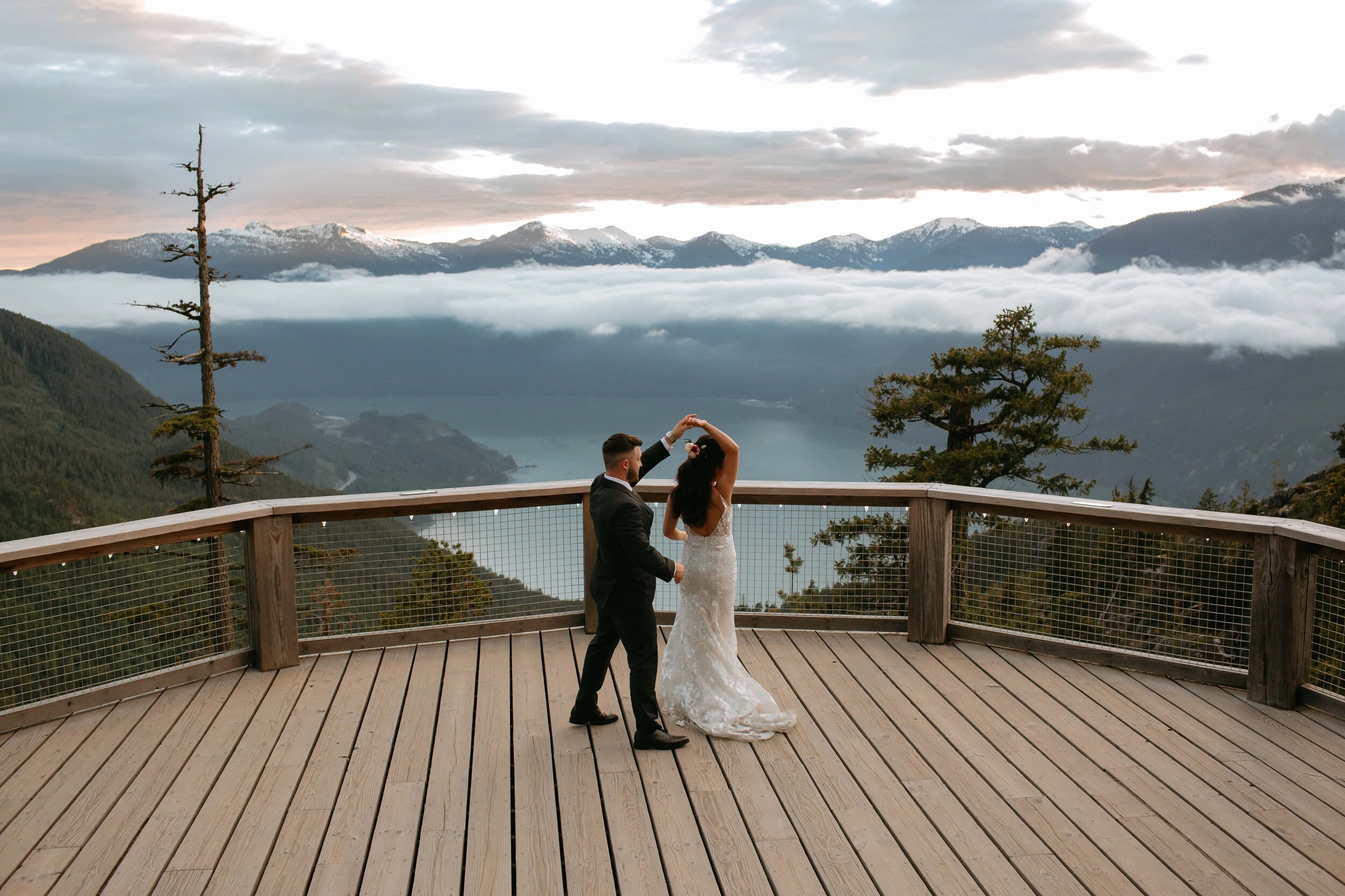 A bride and groom dancing on a wooden deck with a mountain lake and fog-covered mountains in the background amid a cloudy sky.