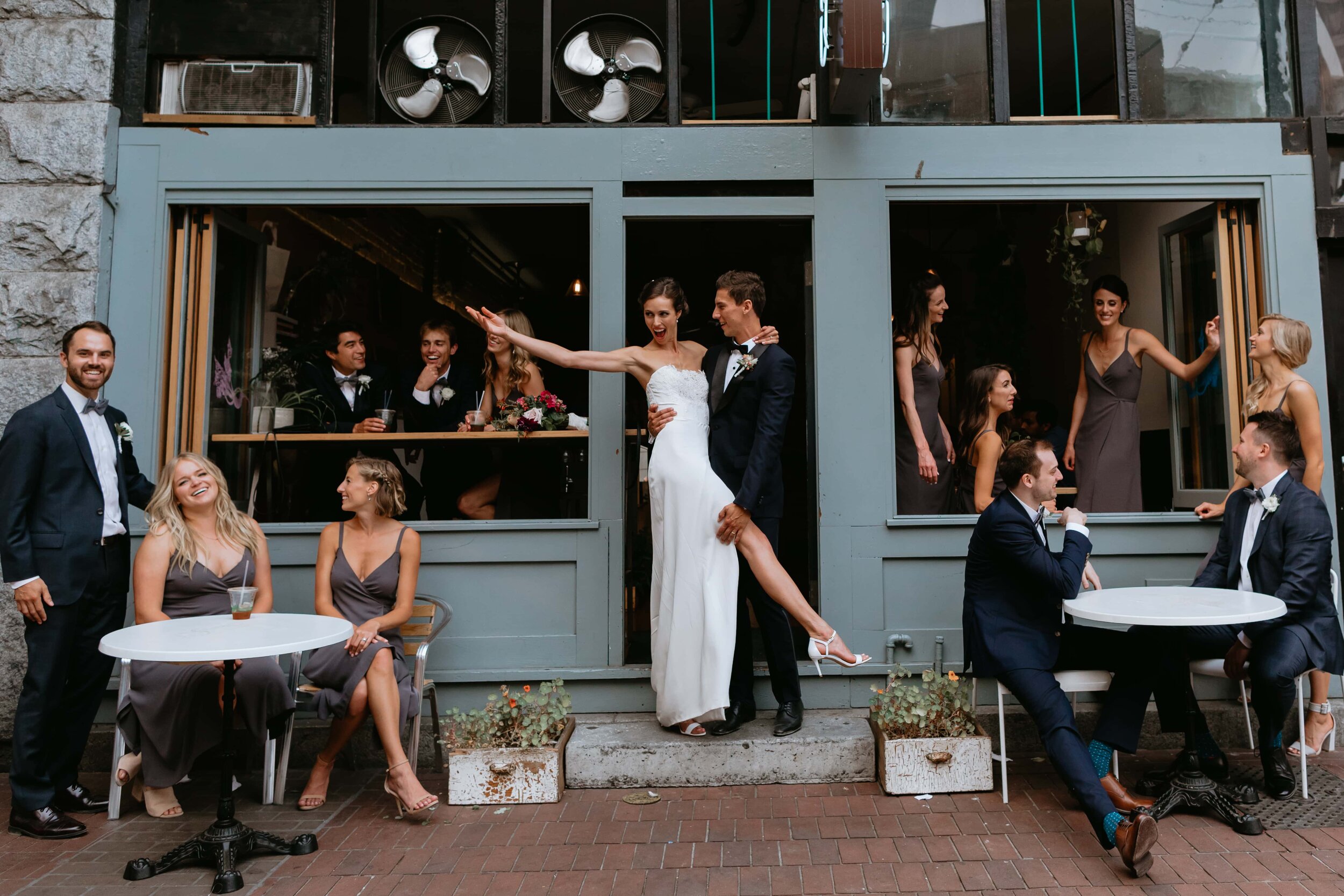 A group of people at a wedding celebration outside a restaurant, with the bride and groom embracing in the center, smiling and enjoying their special day.