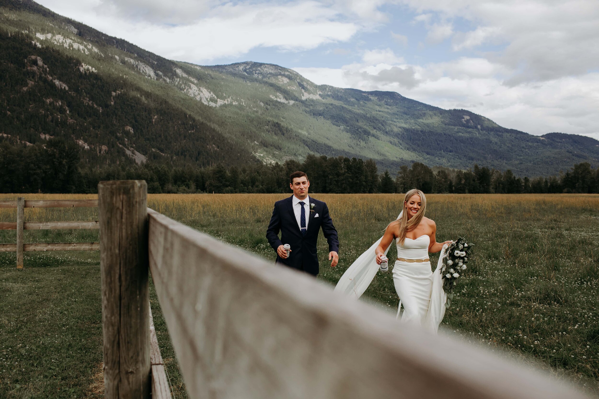 A bride and groom walking through a grassy field with mountains in the background, holding beverages, smiling and enjoying their day.