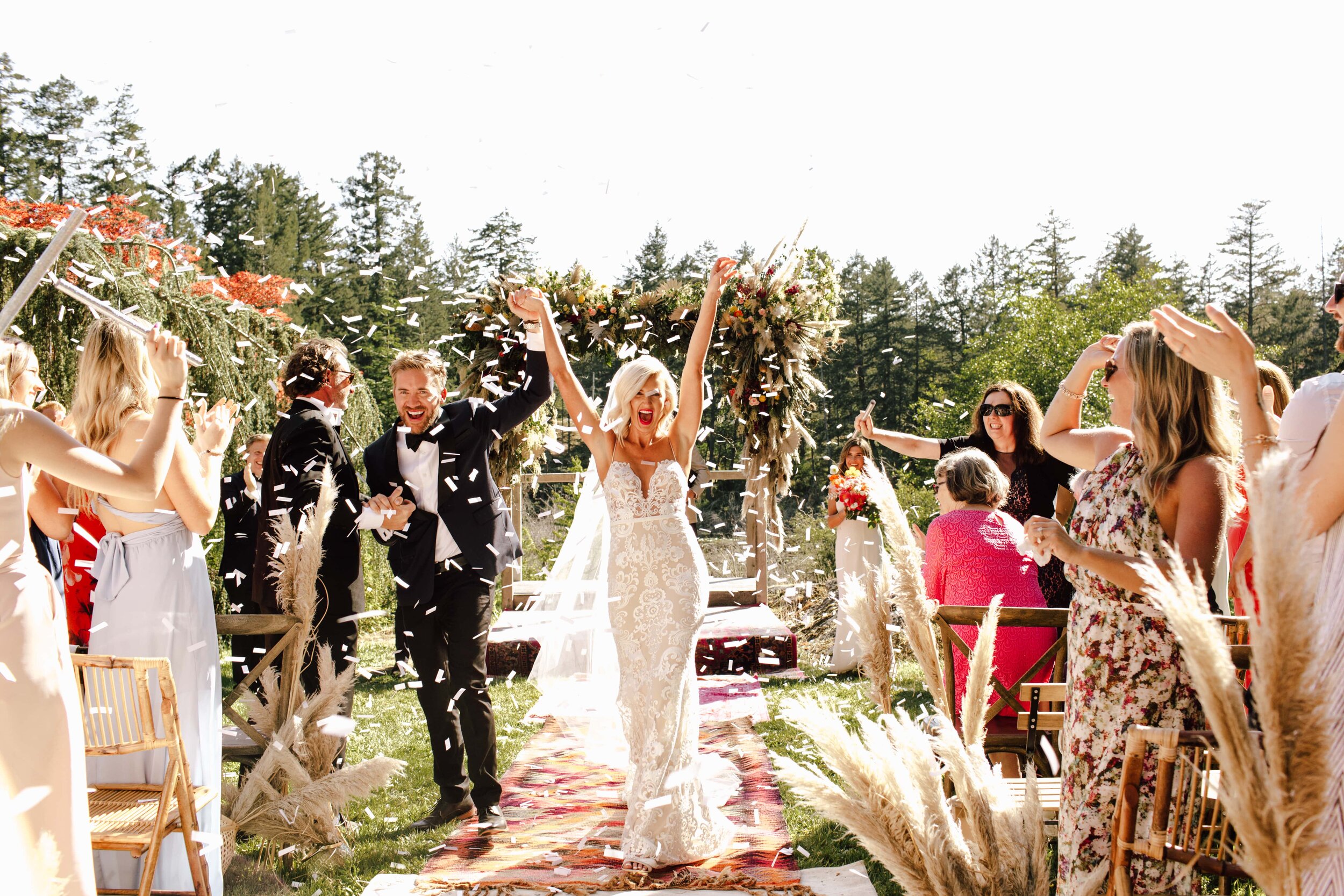 A bride and groom celebrating their wedding as guests throw confetti outdoors on a sunny day.