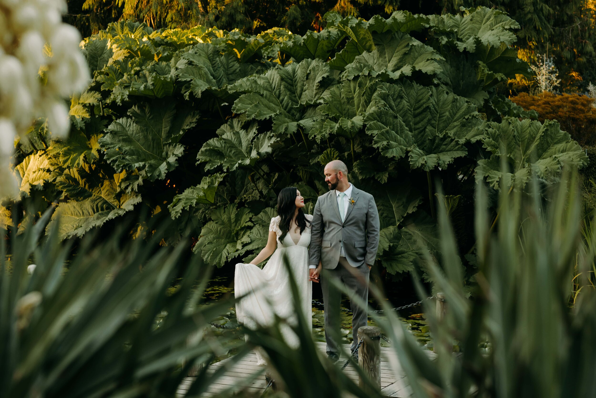 A bride and groom holding hands and gazing at each other on a wooden bridge surrounded by large green leaves and garden foliage.