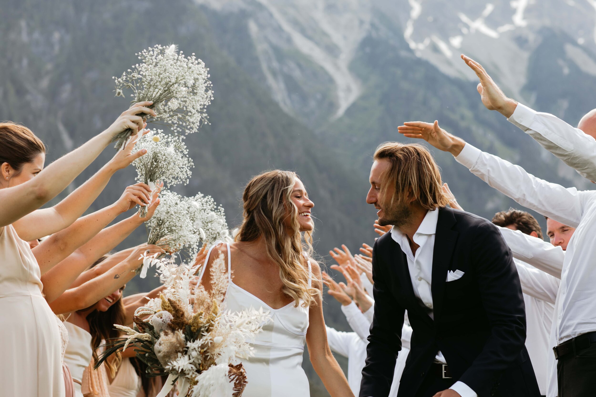 Bride and groom smiling at each other during their wedding while surrounded by wedding guests holding bouquets and raising their hands in celebration, with mountains in the background.