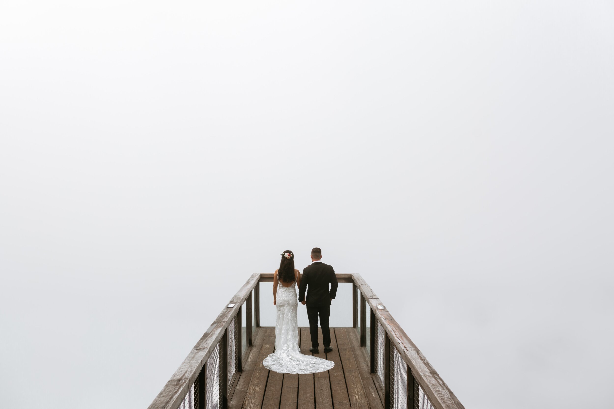 Bride and groom holding hands, walking on a wooden pier into a blank, foggy sky.