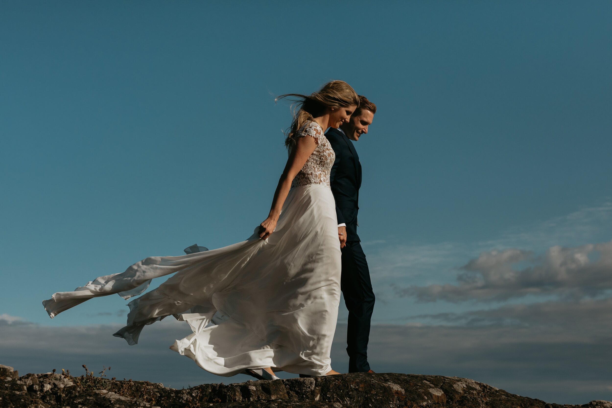 A bride and groom walking on a rocky surface outdoors under a blue sky with scattered clouds, with the bride holding her flowing wedding dress.