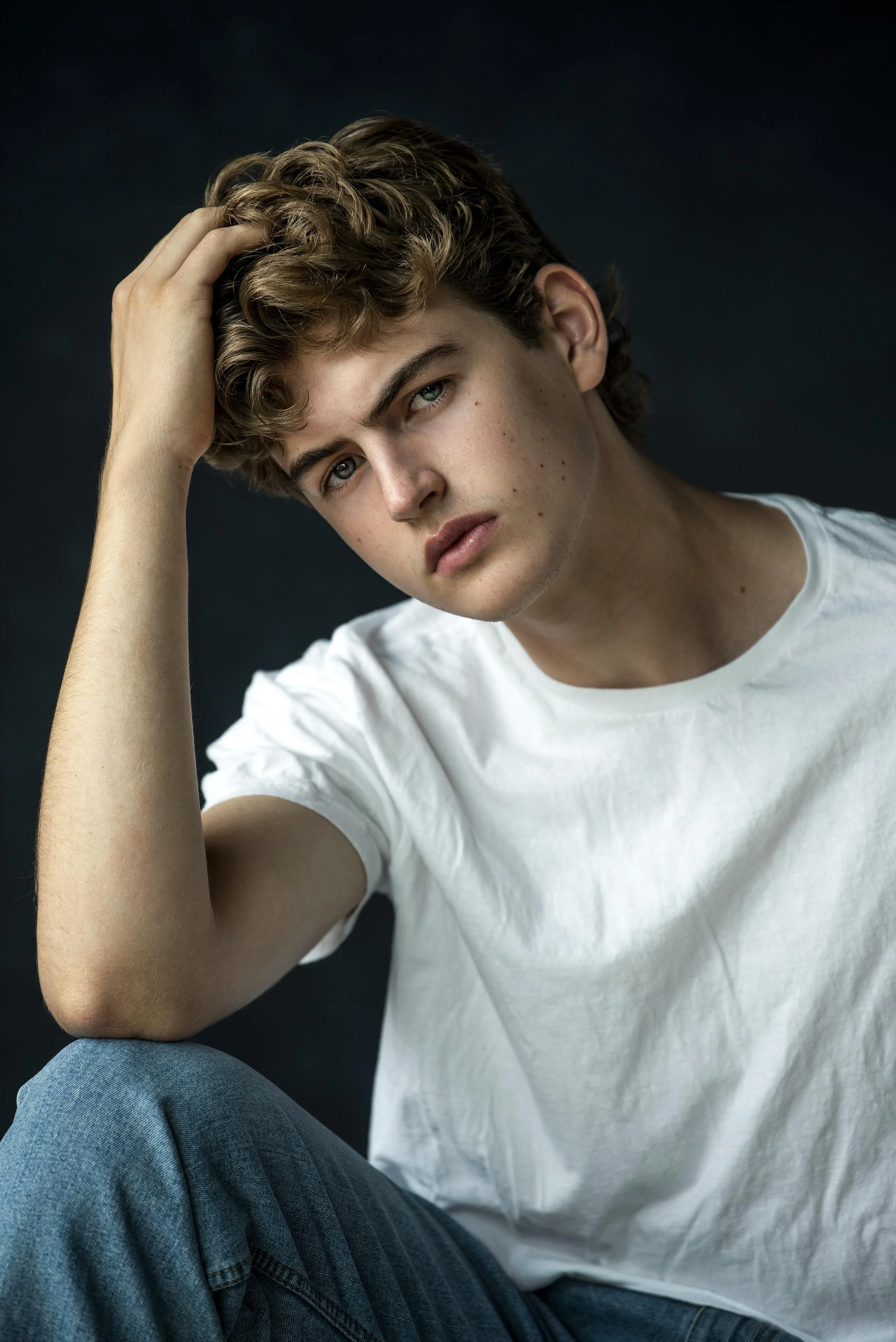 A young man with curly brown hair, blue eyes, and a fair complexion, wearing a white T-shirt and jeans, resting his head on his left hand against a dark background.
