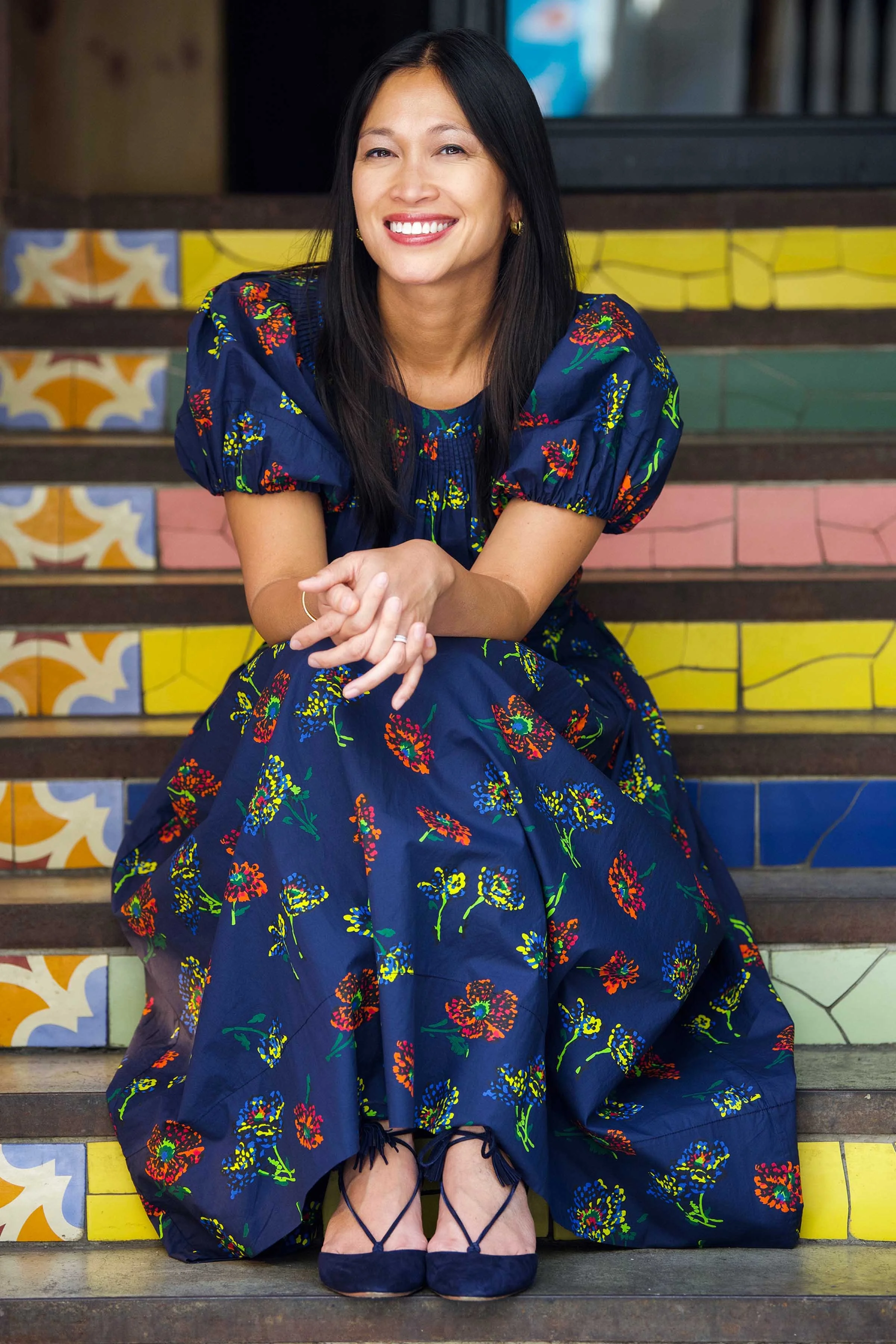 A woman with long dark hair smiling, sitting on colorful tiled stairs, wearing a blue dress with a floral pattern and matching blue heels.