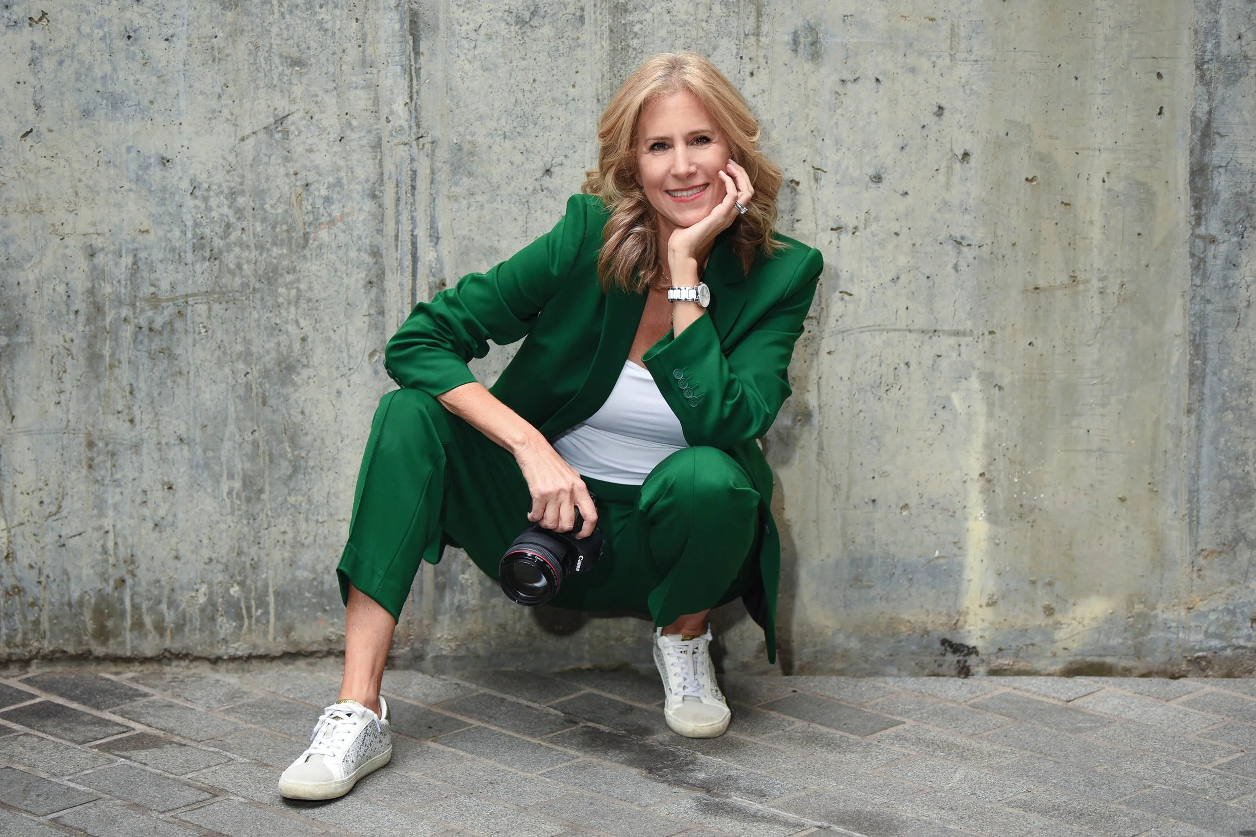 A woman with blond hair sitting against a concrete wall, wearing a green suit, white sneakers, holding a camera, smiling, resting her chin on her hand.