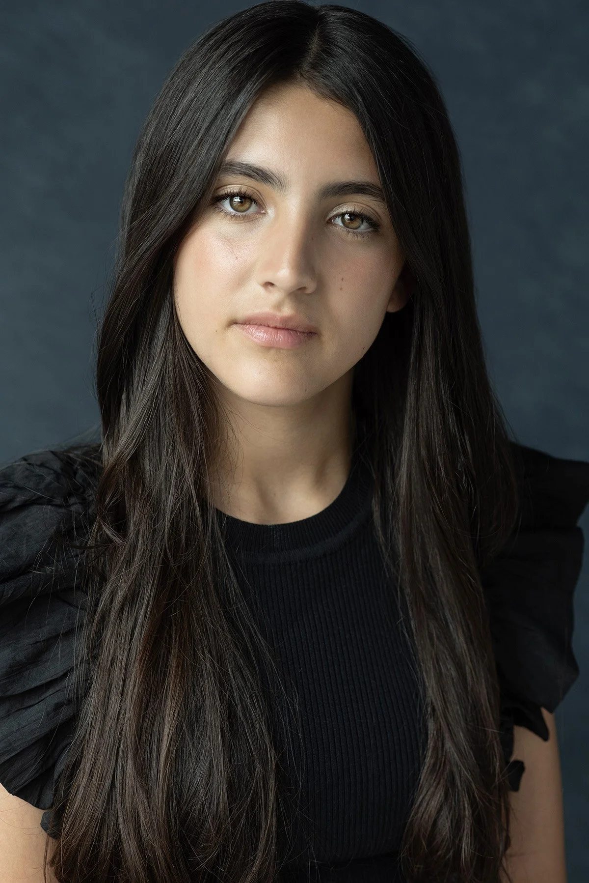 A young woman with long dark hair and hazel eyes, wearing a black top, standing against a dark background.