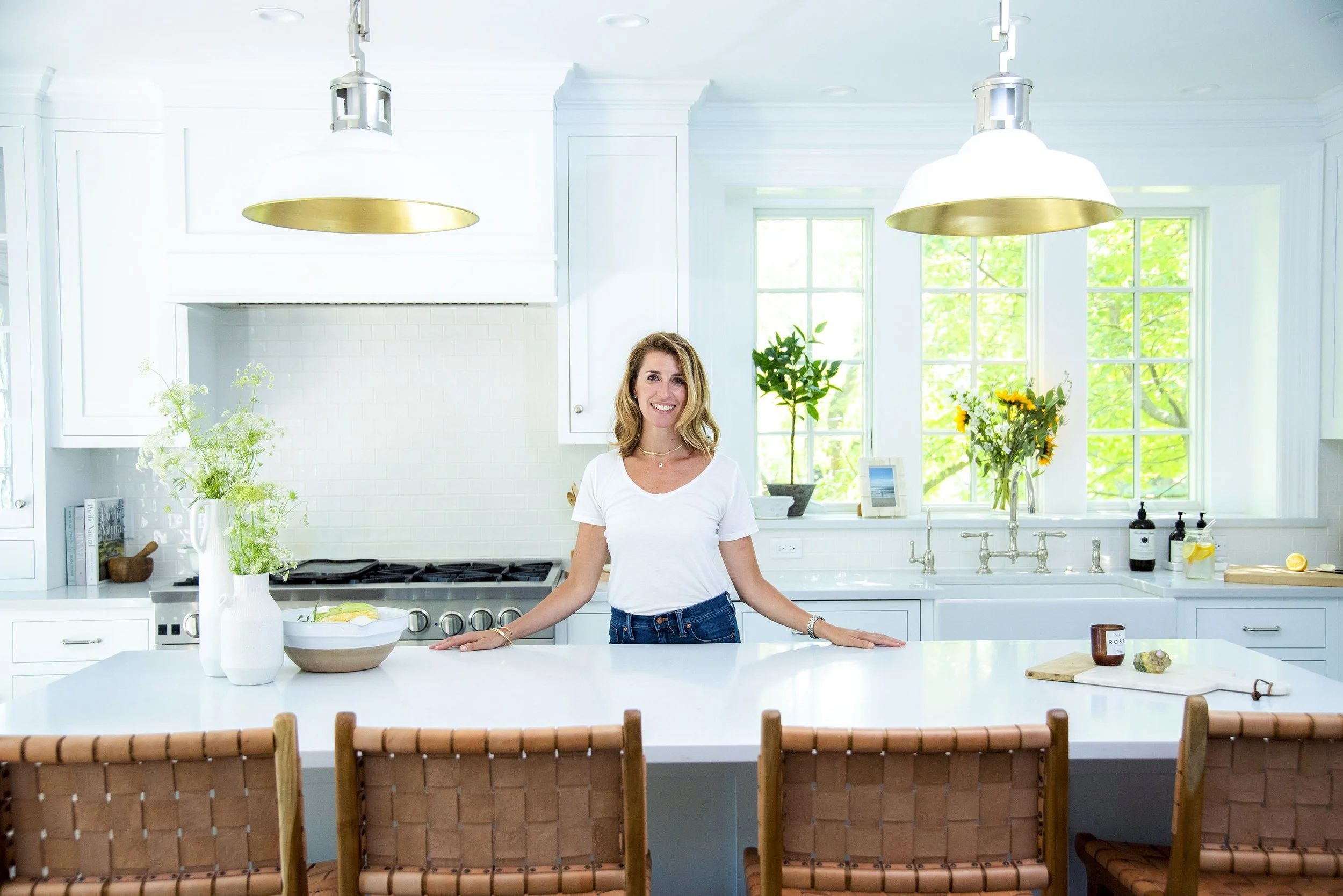 A woman with blond hair smiling in a bright, modern white kitchen with large windows, decorated with green plants and flowers, standing behind a white kitchen island with brown woven chairs in the foreground.
