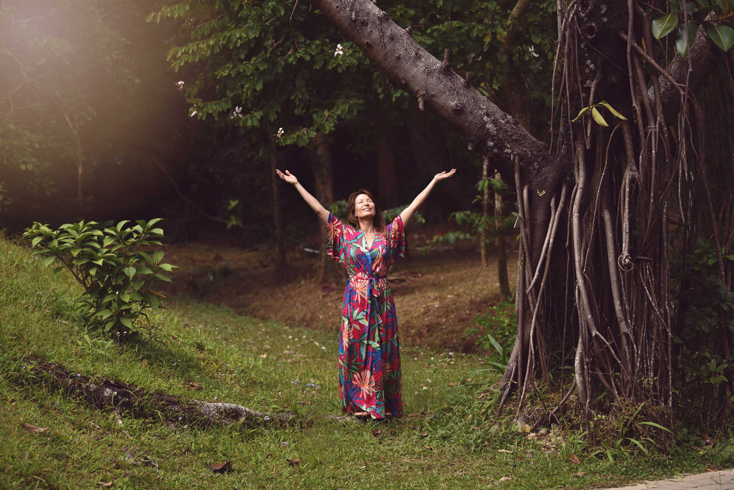 A woman in a colorful dress with arms raised, smiling, standing in a lush garden near a large tree with sprawling roots and green foliage.