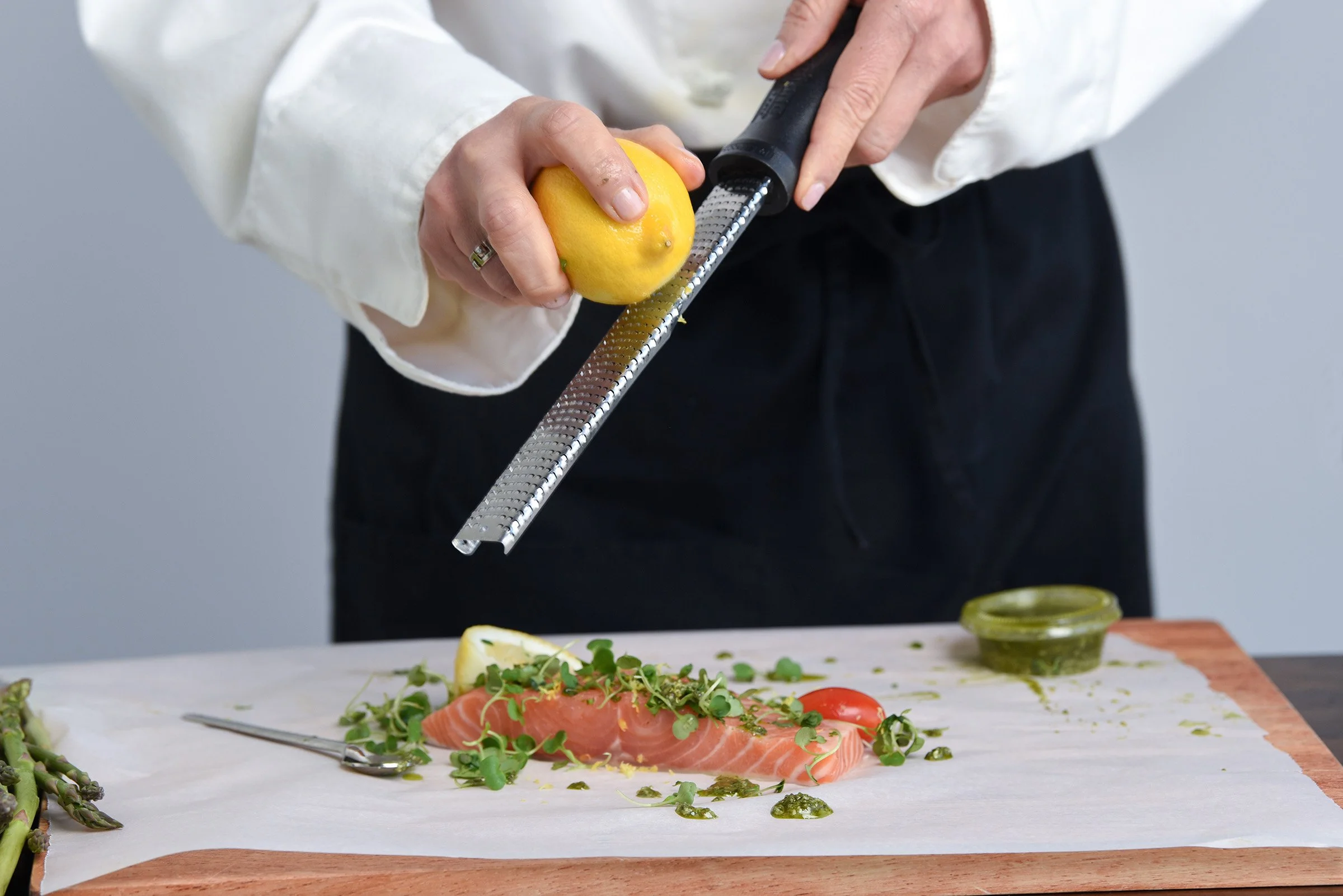 Person garnishing sliced salmon with herbs using a microplane grater on a kitchen counter, with fresh vegetables and dipping sauce nearby.