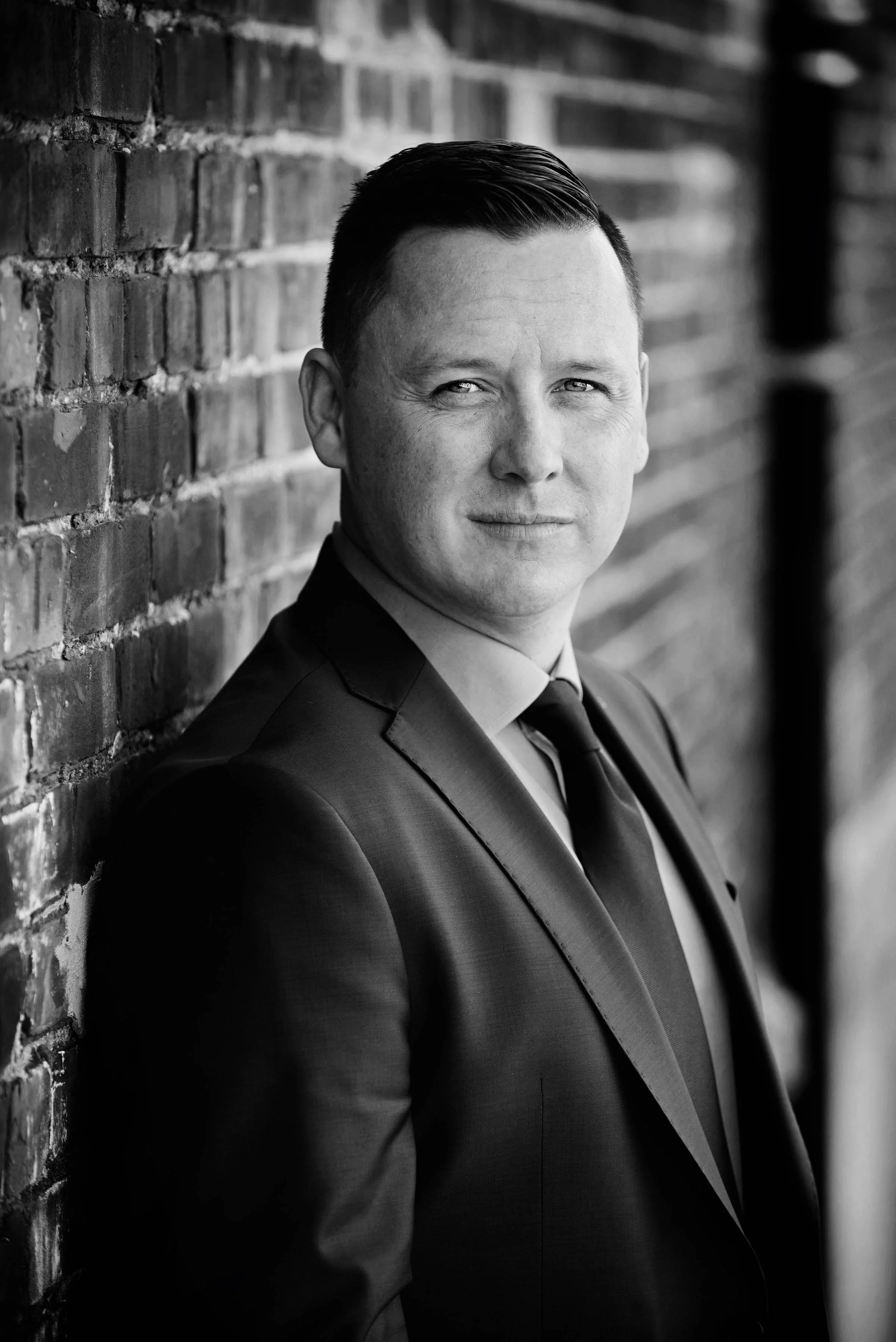 Black and white portrait of a man in a suit leaning against a brick wall.