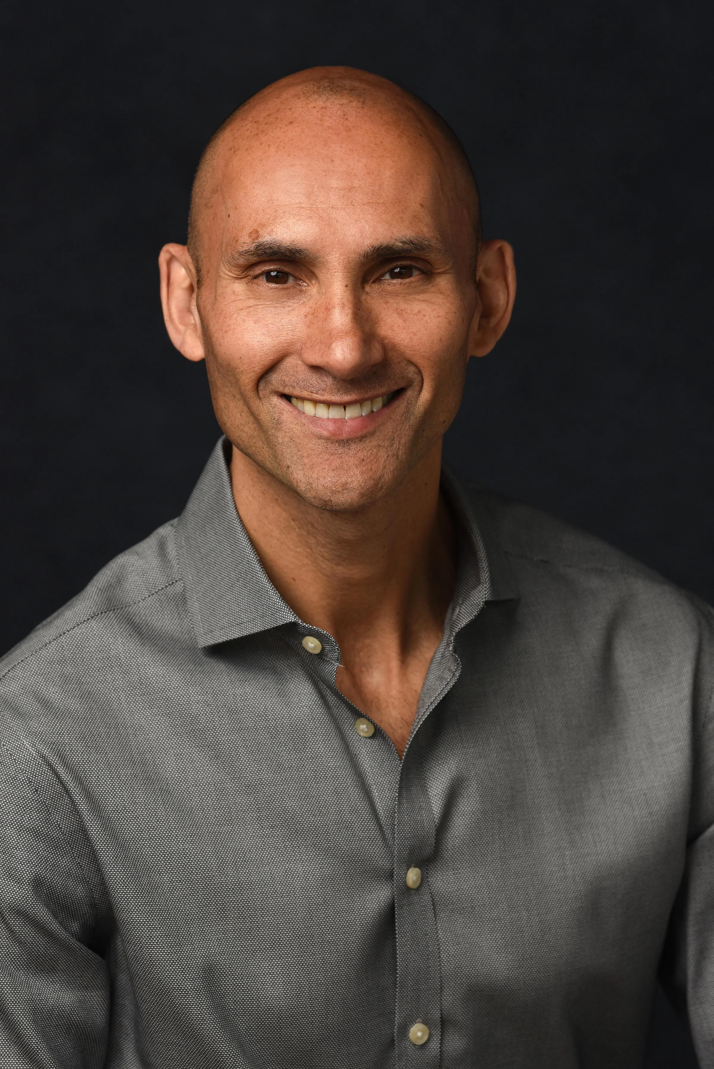 A smiling man with a shaved head wearing a grey dress shirt against a black background.