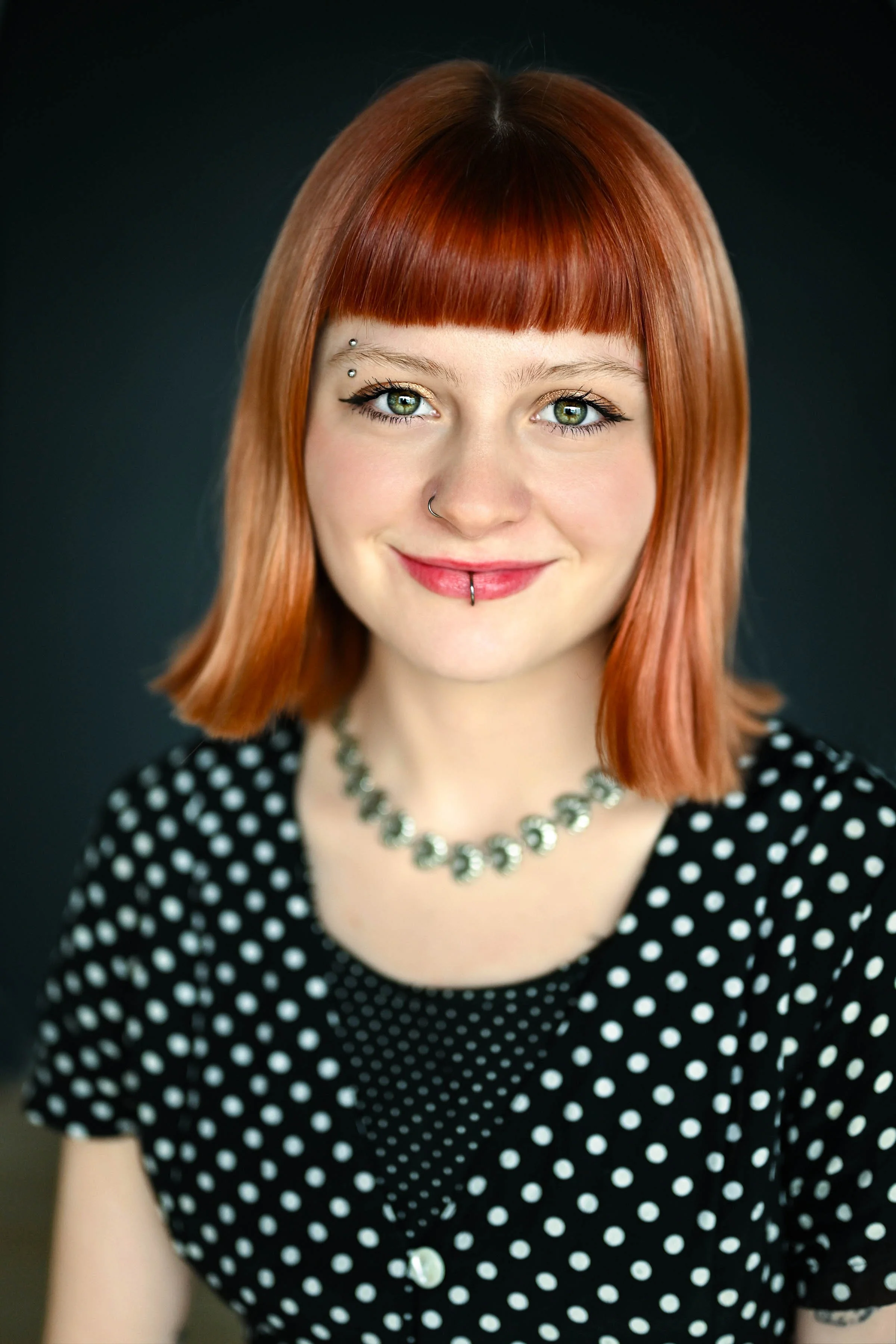 A young woman with red hair, green eyes, and facial piercings, wearing a black and white polka dot top and a silver necklace, smiling against a dark background.