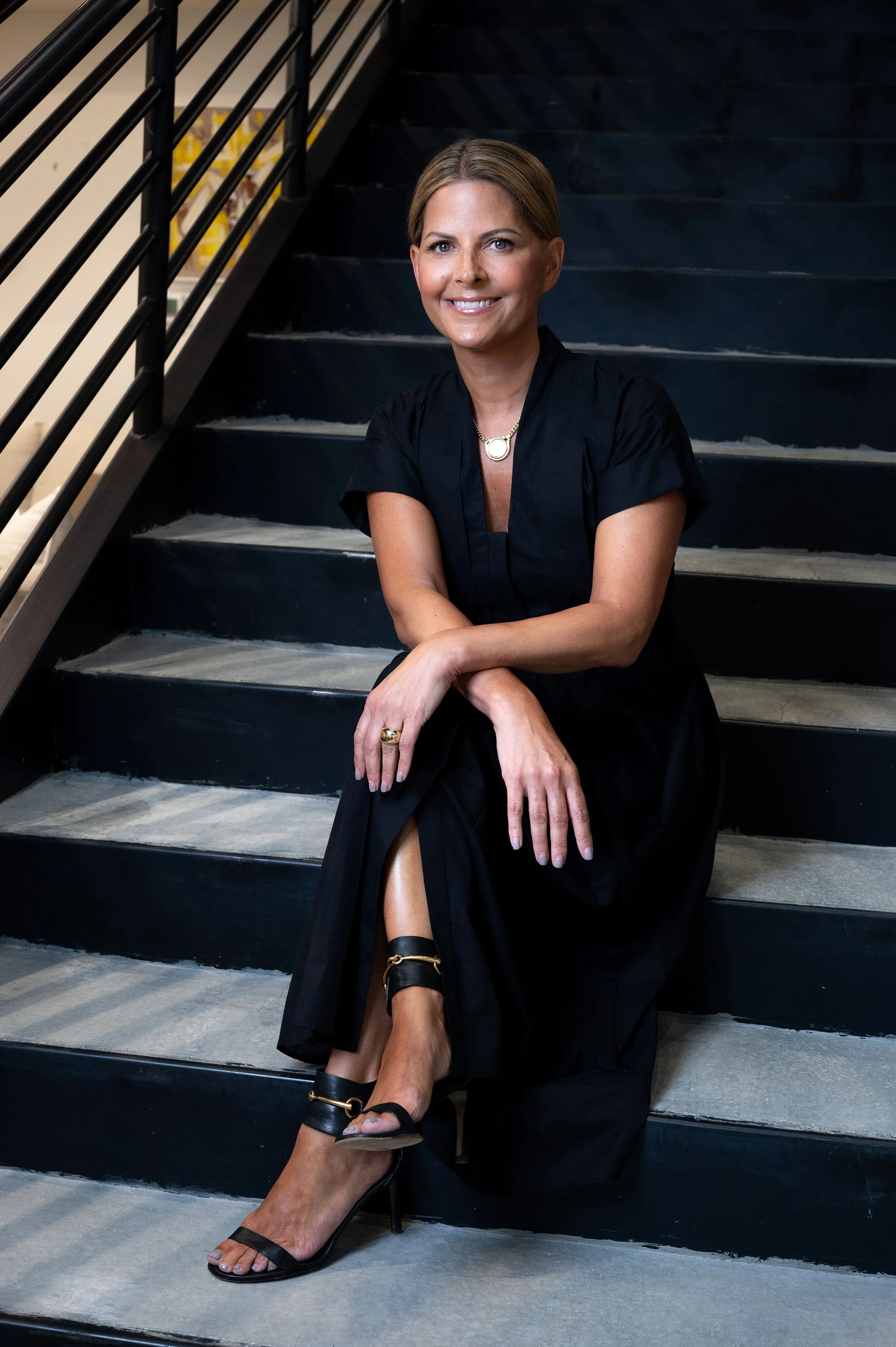 A woman sitting on black staircase stairs, wearing a black dress and black high-heeled sandals, smiling, with her legs crossed, and a gold ring and necklace.