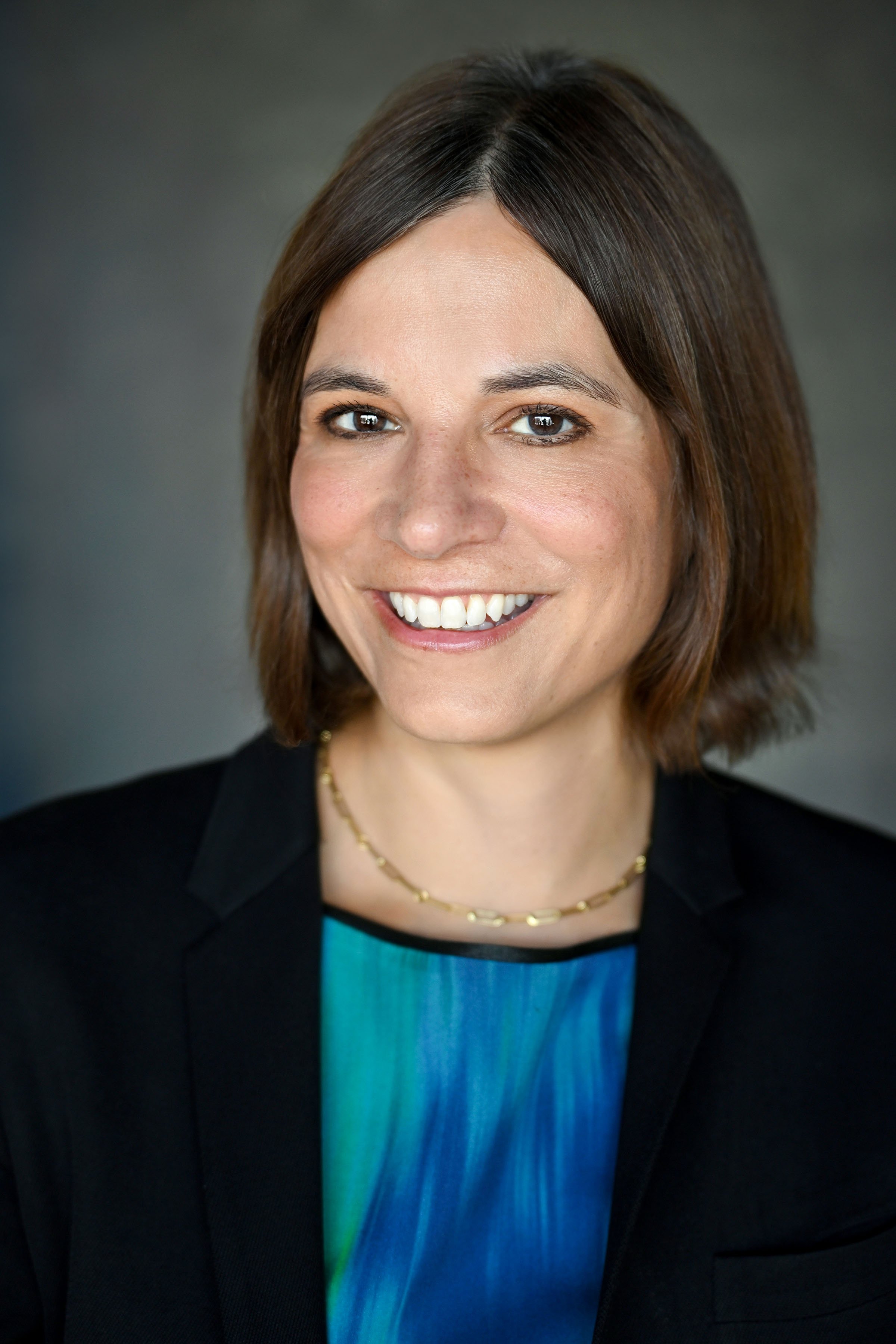 Professional headshot of a woman with short brown hair, wearing a black blazer and a blue top, smiling against a dark grey background.