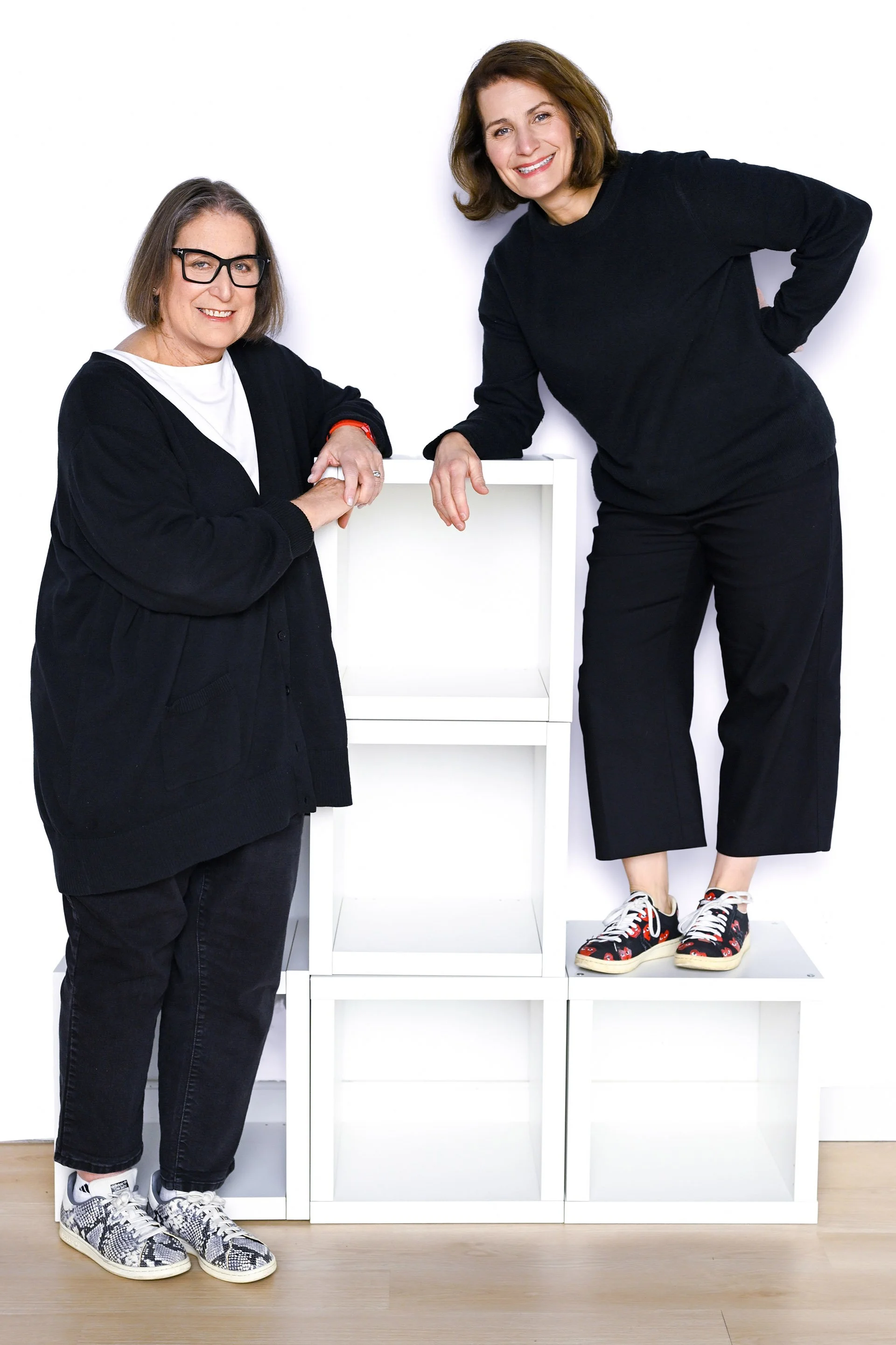 Two women smiling, one with glasses and a black hoodie, the other standing on a white shelf wearing sneakers with a black hoodie, with a white background and empty white shelves.